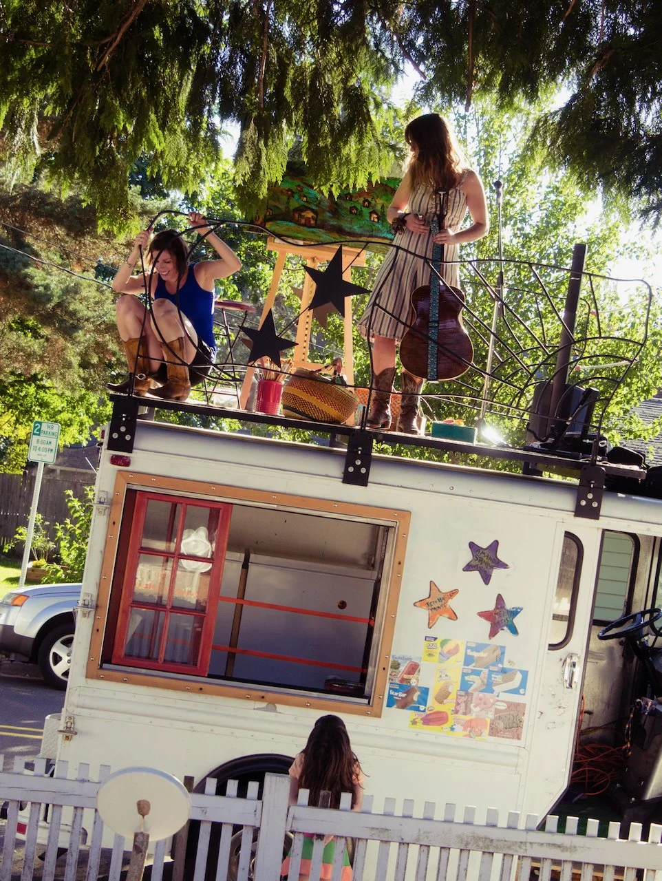 Two young women on the roof of a decorated mobile structure, one with a guitar and the other adjusting a wire, with a girl looking over the fence in front. The scene is outdoors with trees overhead and cars in the background.