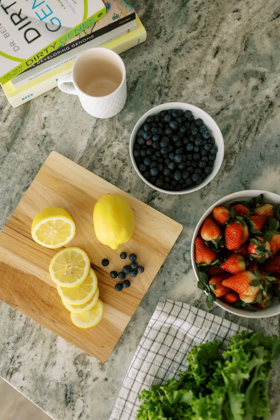 A kitchen counter with a cutting board of lemon slices, blueberries, and a lemon, a bowl of strawberries, a bowl of blueberries, a white mug, a stack of books, and a bowl of strawberries, with a kitchen towel and some leafy greens.