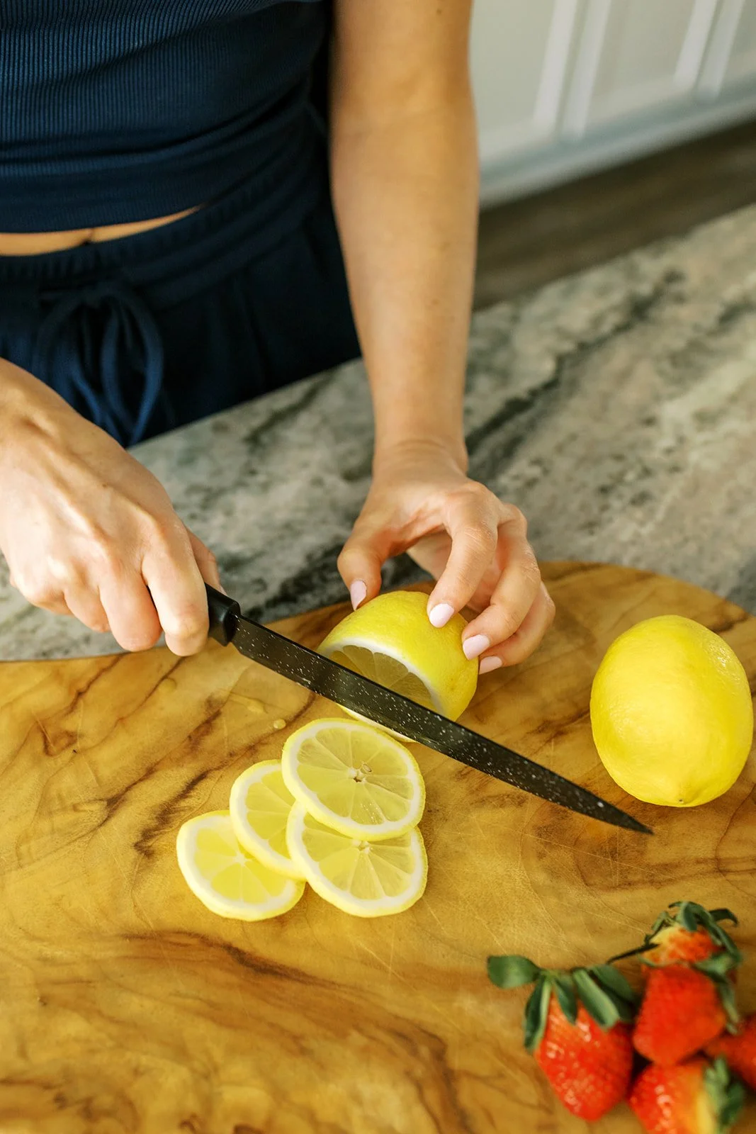 A person is slicing a lemon on a wooden cutting board with a black knife. There are several lemon slices already cut on the board. A whole lemon and some strawberries are also visible on the board.