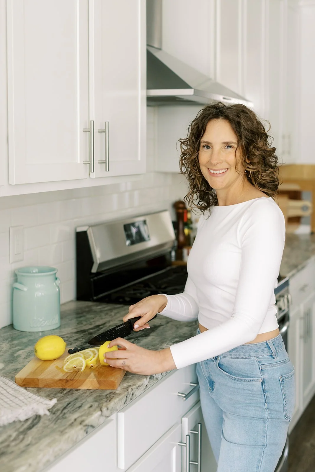 A woman with curly brown hair wearing a white long-sleeve shirt and light blue jeans is cutting lemons on a wooden cutting board in a modern kitchen.