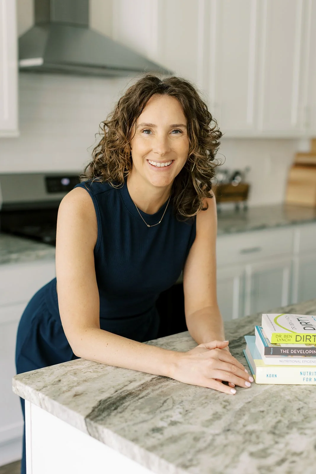 A smiling woman with curly brown hair, wearing a navy sleeveless dress, standing in a kitchen with a stack of books on a marble countertop.