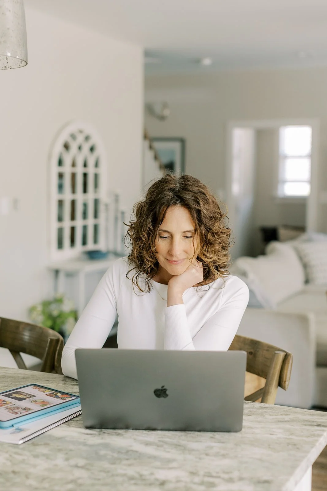 Woman with curly hair sitting at a kitchen table working on a MacBook.