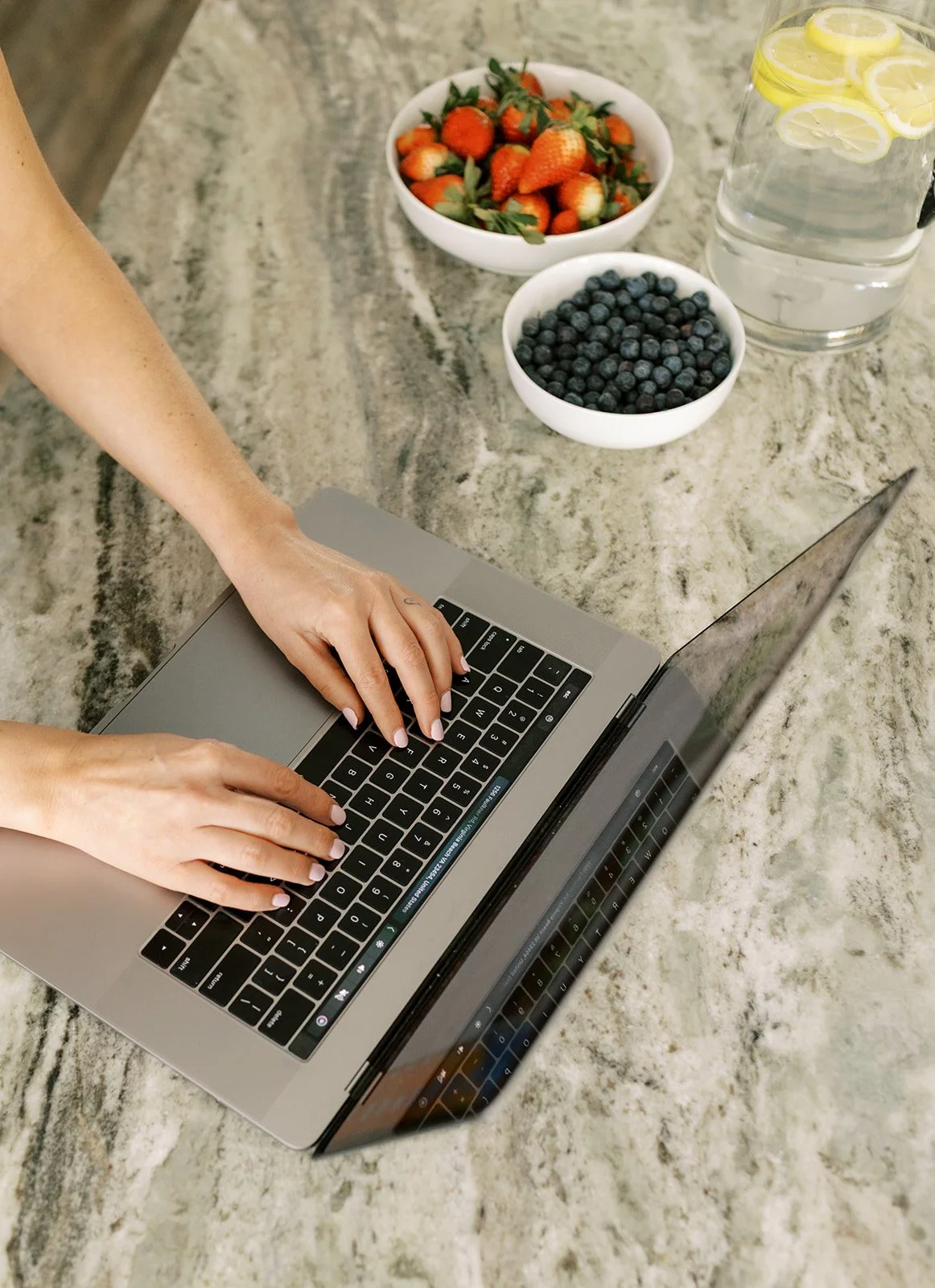 Hands typing on a silver laptop on a marble countertop, with bowls of strawberries and blueberries, a pitcher of lemon water, and a glass of water with lemon slices.