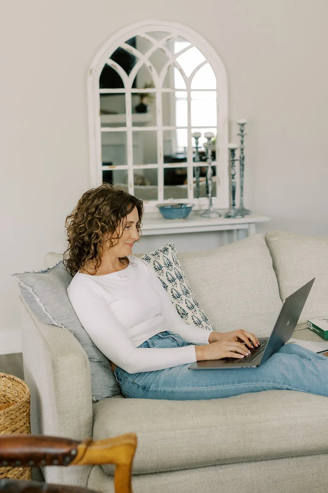 Woman sitting on a light-colored sofa working on a laptop in a bright living room with a decorative mirror on the wall behind her.