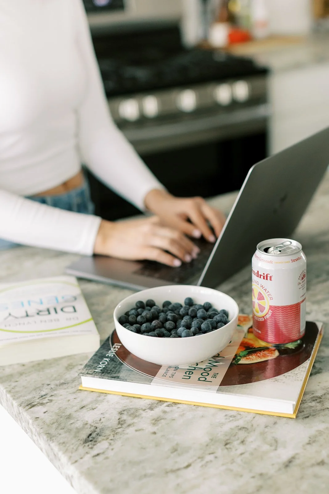 A person in a white long sleeve shirt and jeans typing on a silver laptop on a kitchen counter. On the counter, there's a bowl of blueberries, a can of pink lemonade, a book, and a magazine.