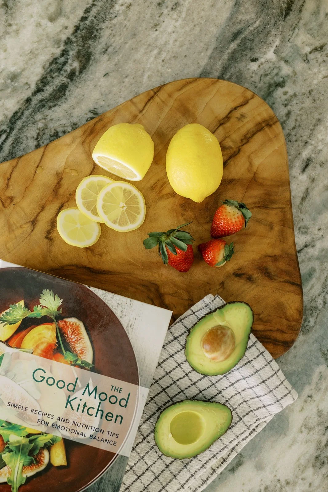 A wooden cutting board with sliced lemons, whole lemons, strawberries, and halved avocado on a marble countertop with a kitchen book.