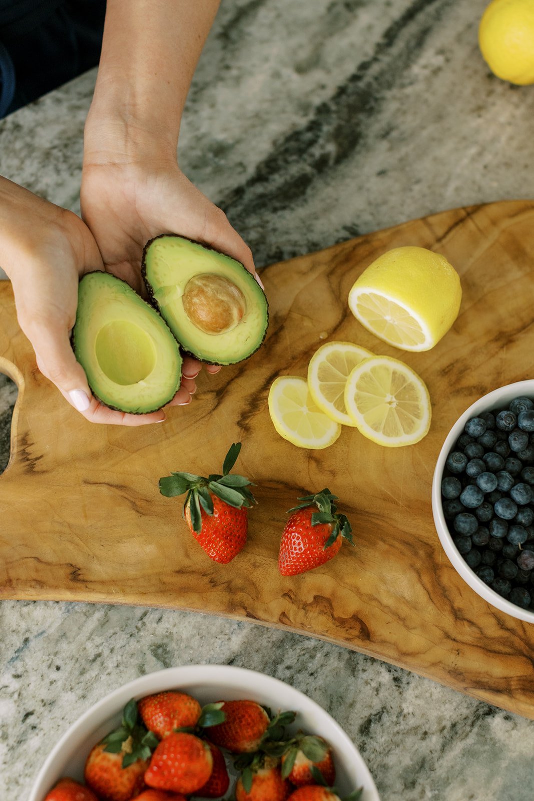 Hands holding halved avocado on a wooden surface with sliced lemon, blueberries in a bowl, and strawberries nearby.