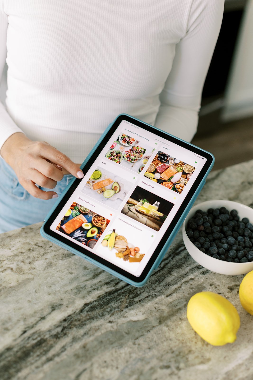 Person in white shirt holding a tablet with a food and nutrition app open in a kitchen, with blueberries and lemons on the marble countertop.