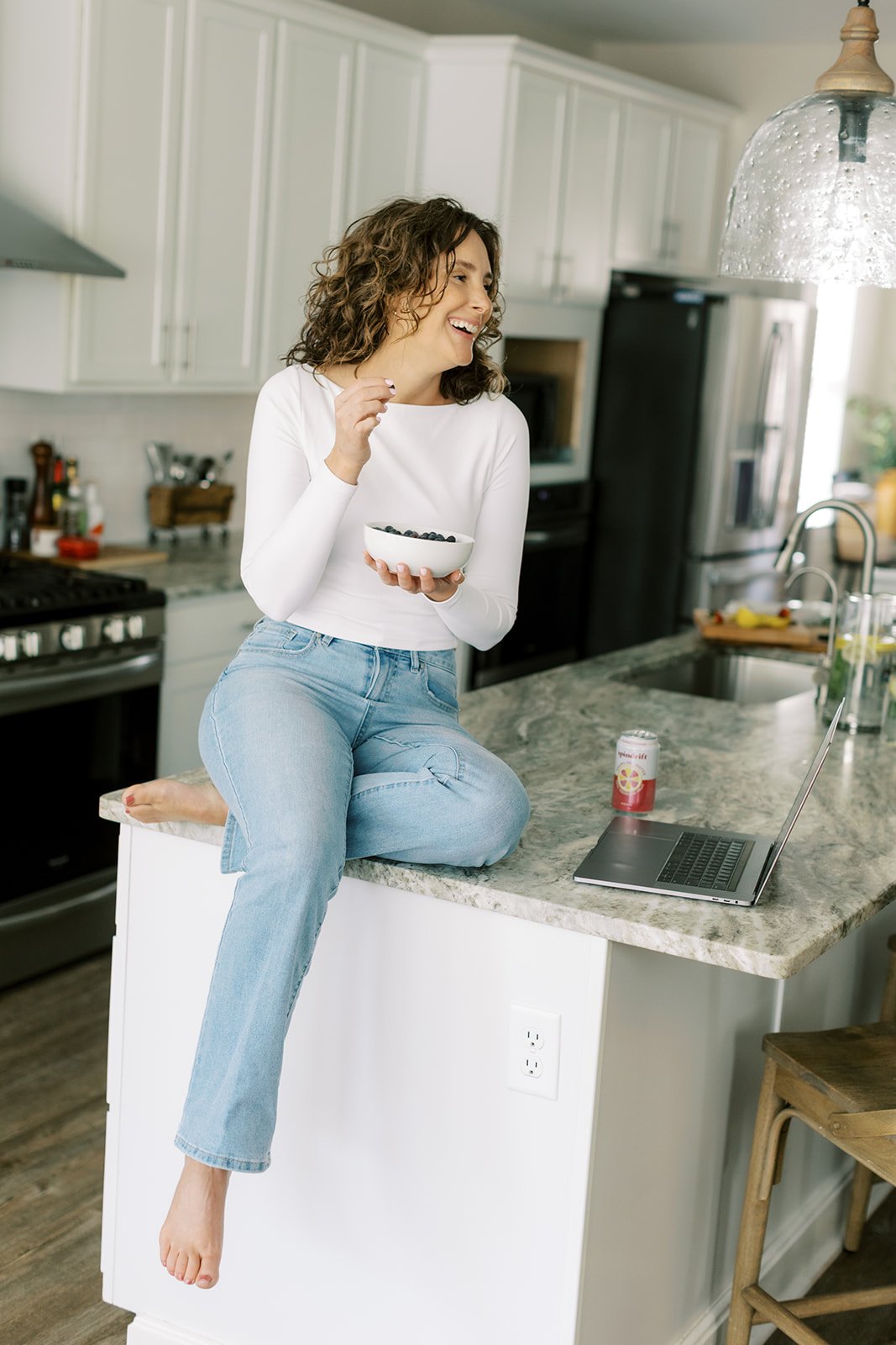 A woman with curly hair in a white long sleeve shirt and light blue jeans sitting on a kitchen counter, laughing while holding a bowl of blueberries, with a laptop and a canned drink nearby.