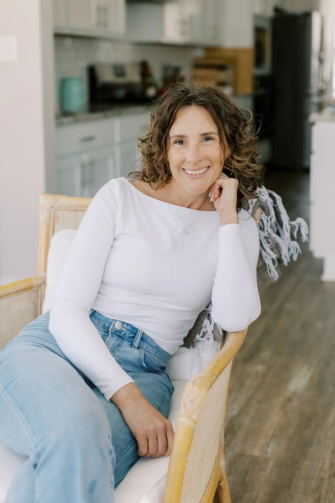 A woman with curly brown hair wearing a white long-sleeve shirt and blue jeans sitting on a yellow wicker chair, smiling at the camera inside a home.
