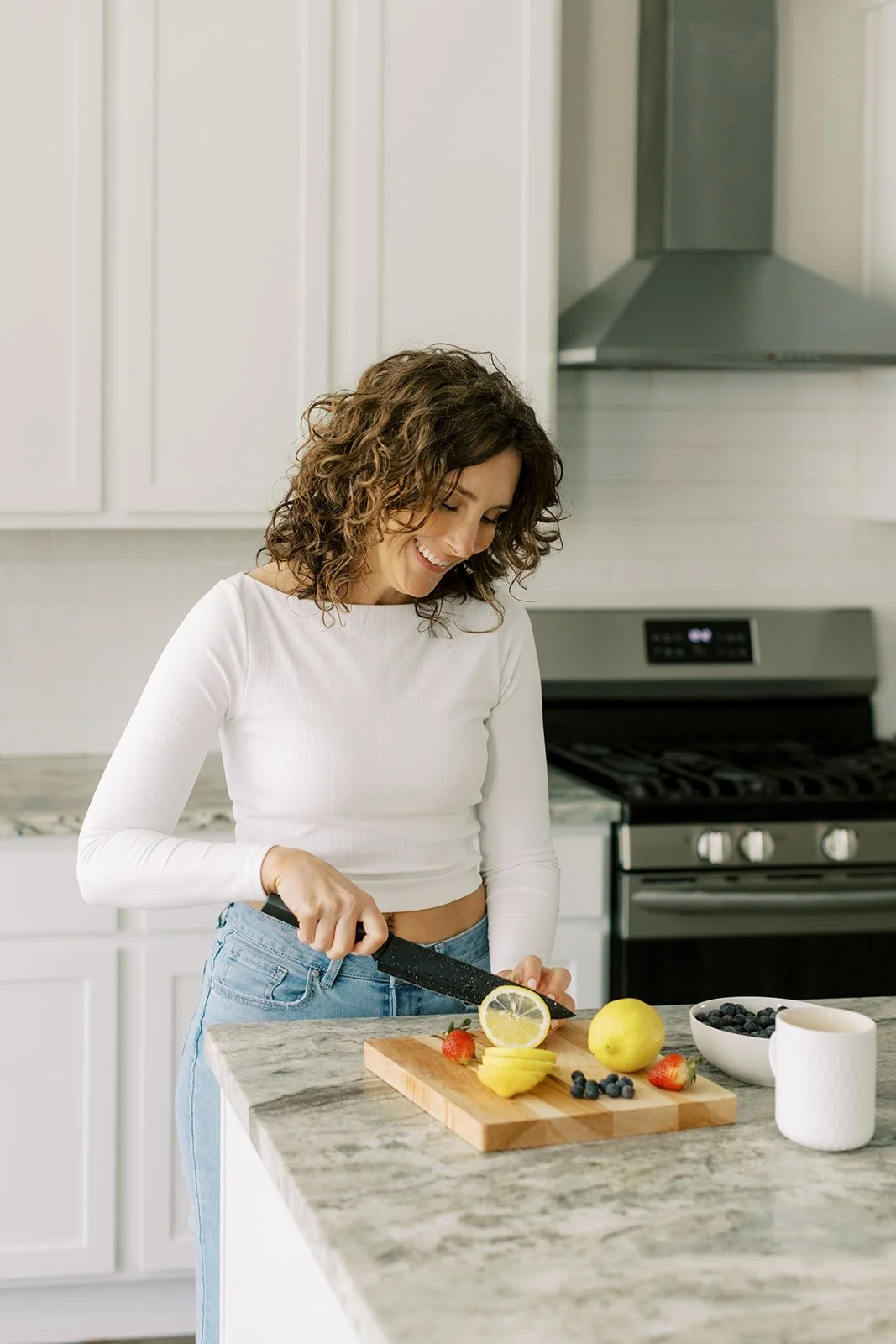 A woman slicing strawberries and lemons on a kitchen counter with a bowl of blueberries and a mug nearby.