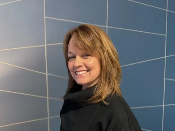 Woman with shoulder-length blonde hair smiling, standing against a blue tiled wall.