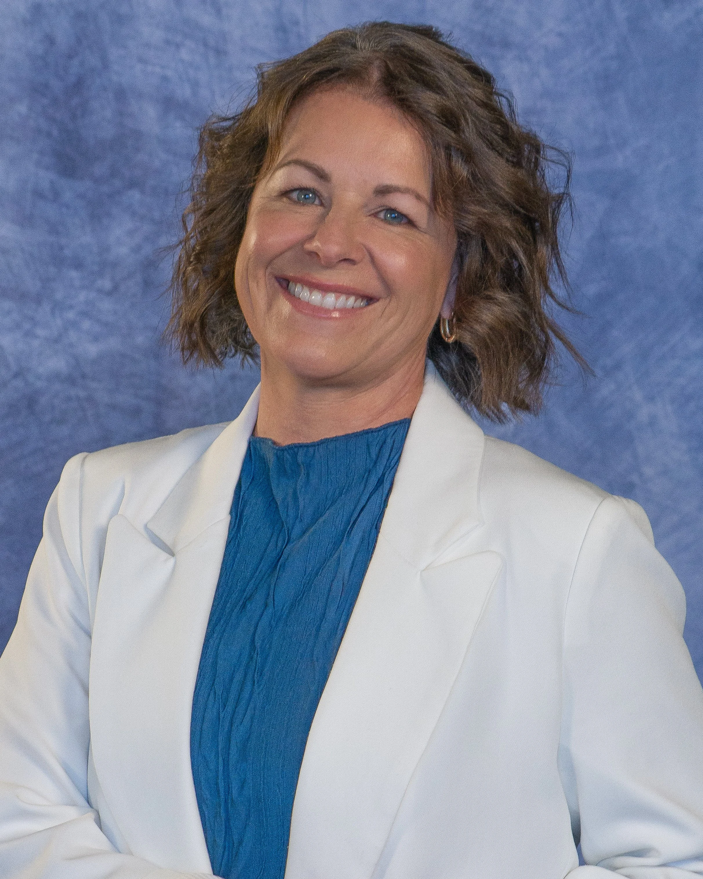 Portrait of a woman with dark wavy hair wearing hoop earrings and a gray turtleneck sweater, smiling against a plain light background.