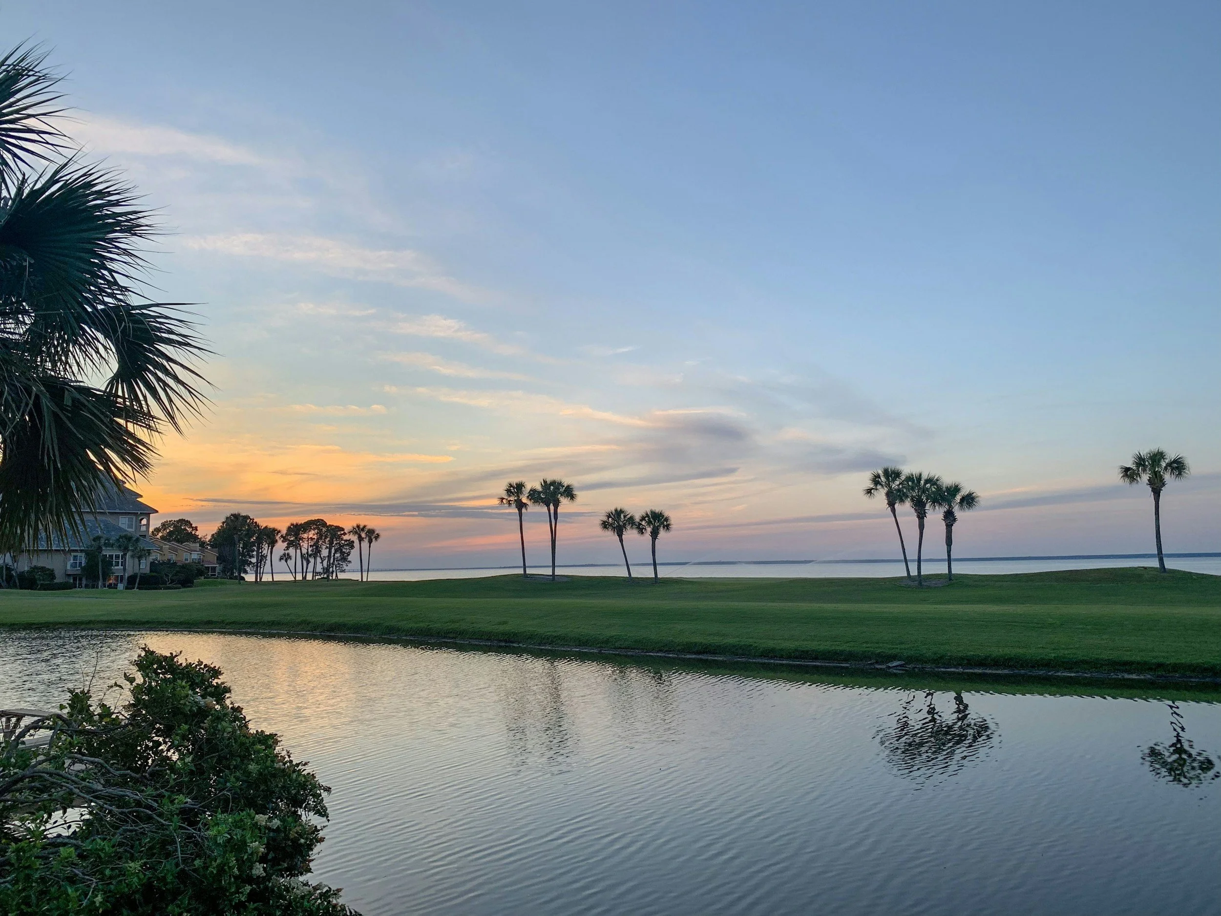 Sunset over a serene water body with palm trees and houses in the distance.