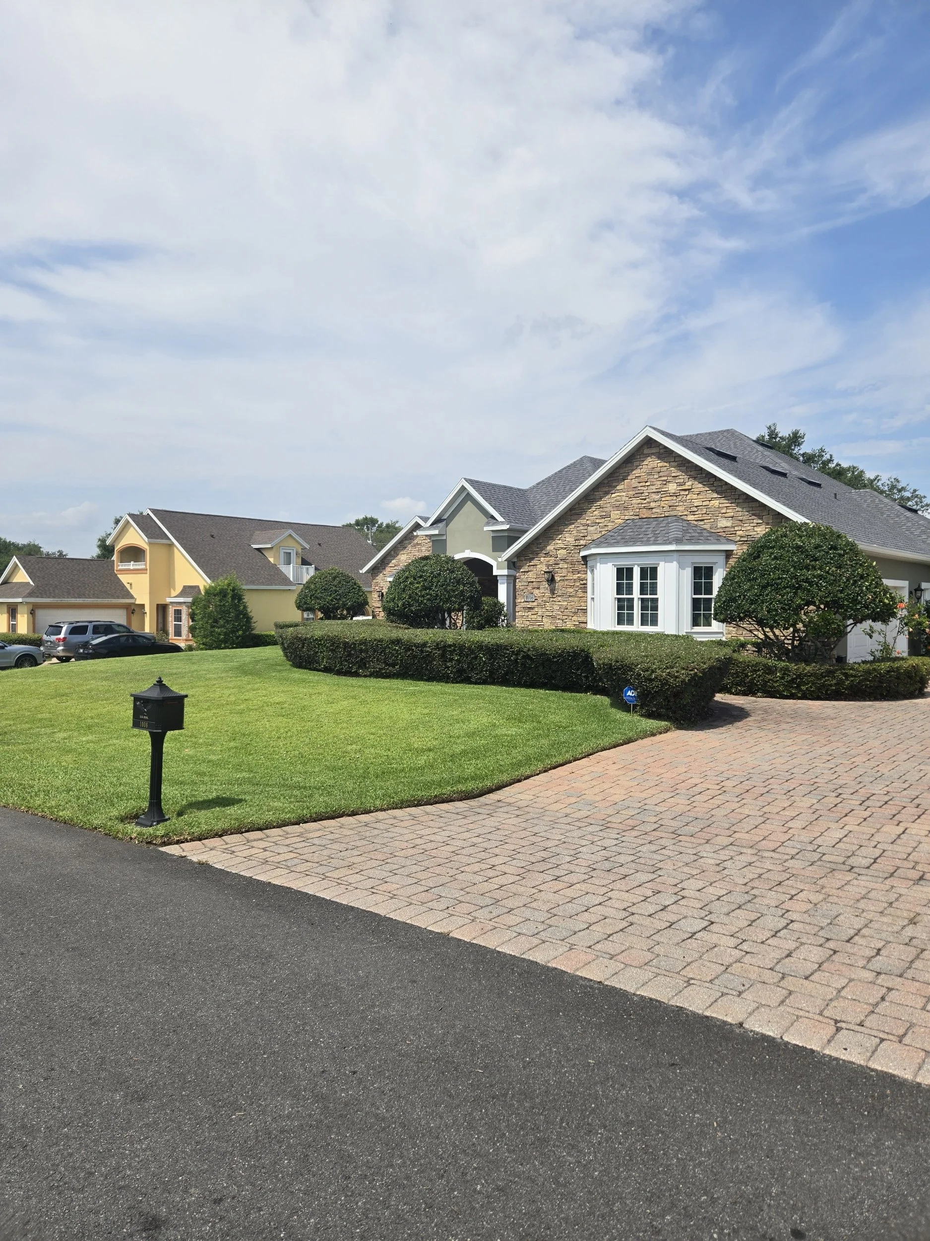 Suburban neighborhood with houses, manicured lawns, a paved driveway, a mailbox, and a partly cloudy sky.