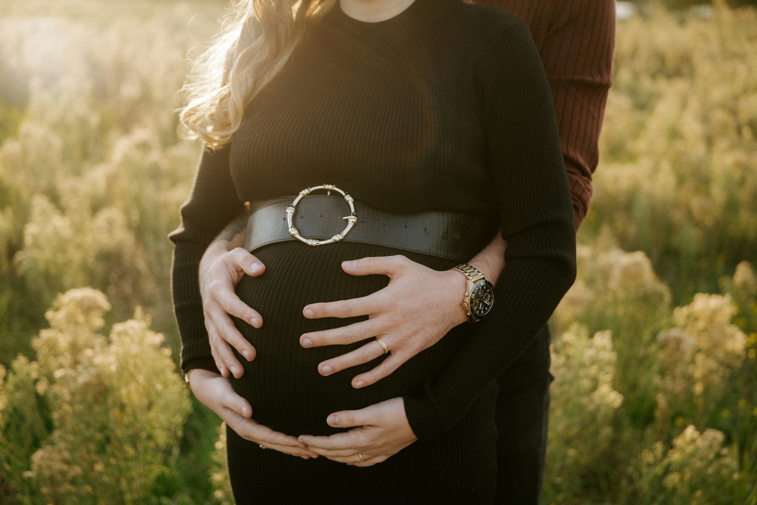 Pregnant woman wearing a black sweater and a wide belt with a large gold buckle, with a man standing behind her, outdoors in a field of yellow flowers, with their hands gently resting on her belly.
