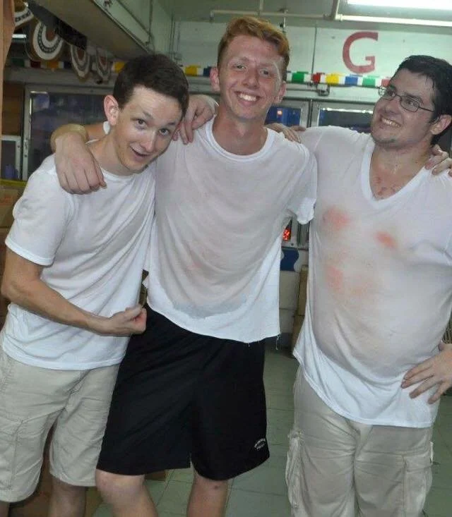 Three young men with light skin and short hair, smiling, with their arms around each other, wearing white t-shirts; one is flexing his arm and the others are standing close together in an indoor setting.