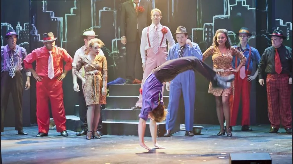 Stage scene from a musical or play with actors dressed in retro clothing, some wearing hats, and a woman in the foreground performing a backbend dance move, with a cityscape backdrop.