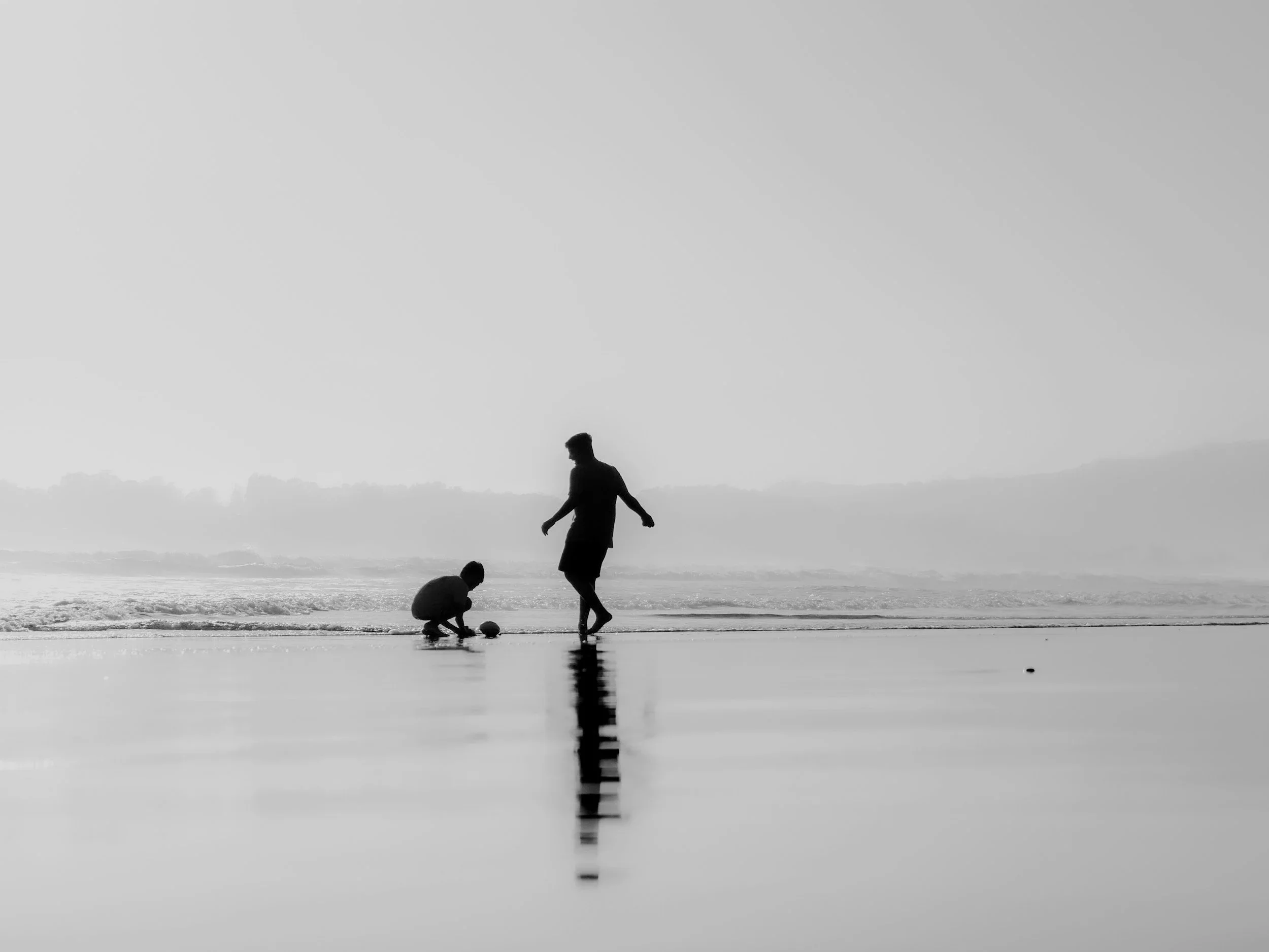 Silhouettes of a man and a child playing with a ball on a beach at sunset or sunrise, reflected in the wet sand.