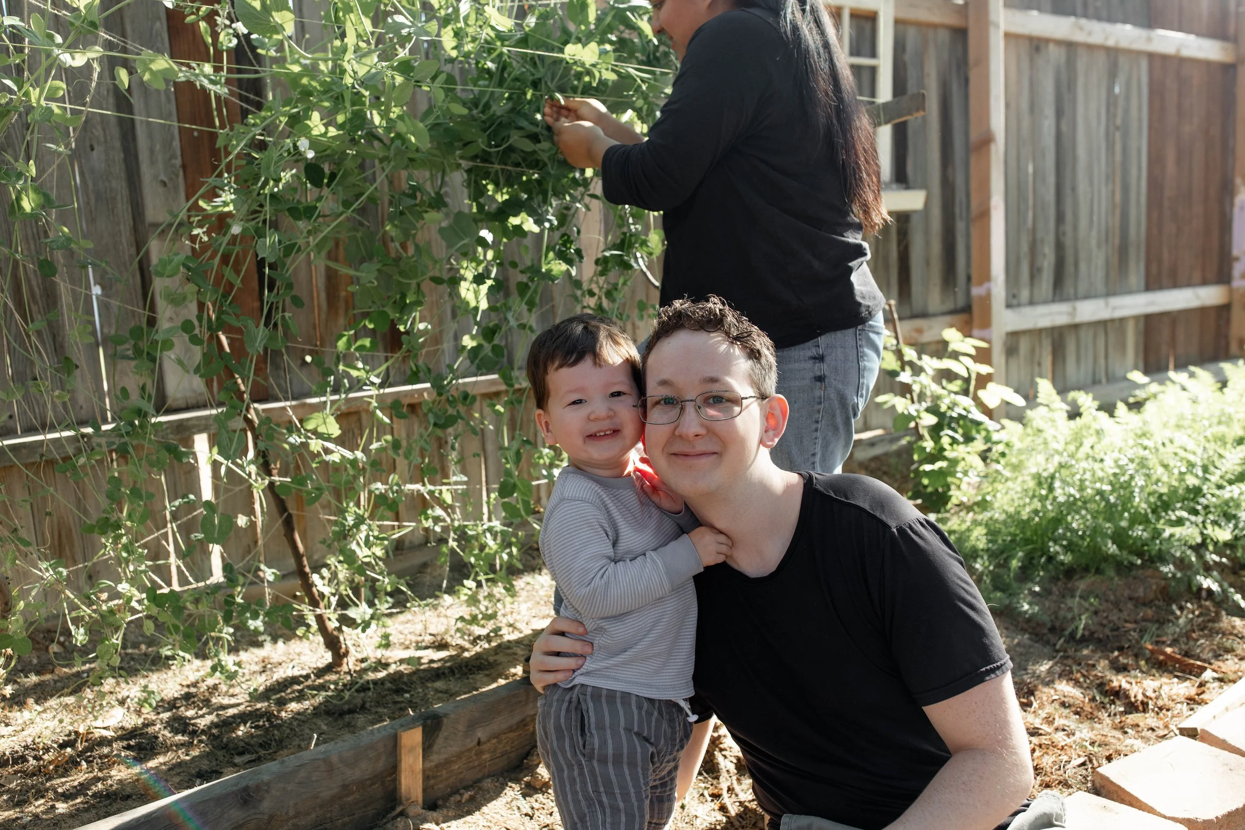 A man and a young boy smiling and hugging in a garden with a wooden fence, plants, and a woman working in the background.