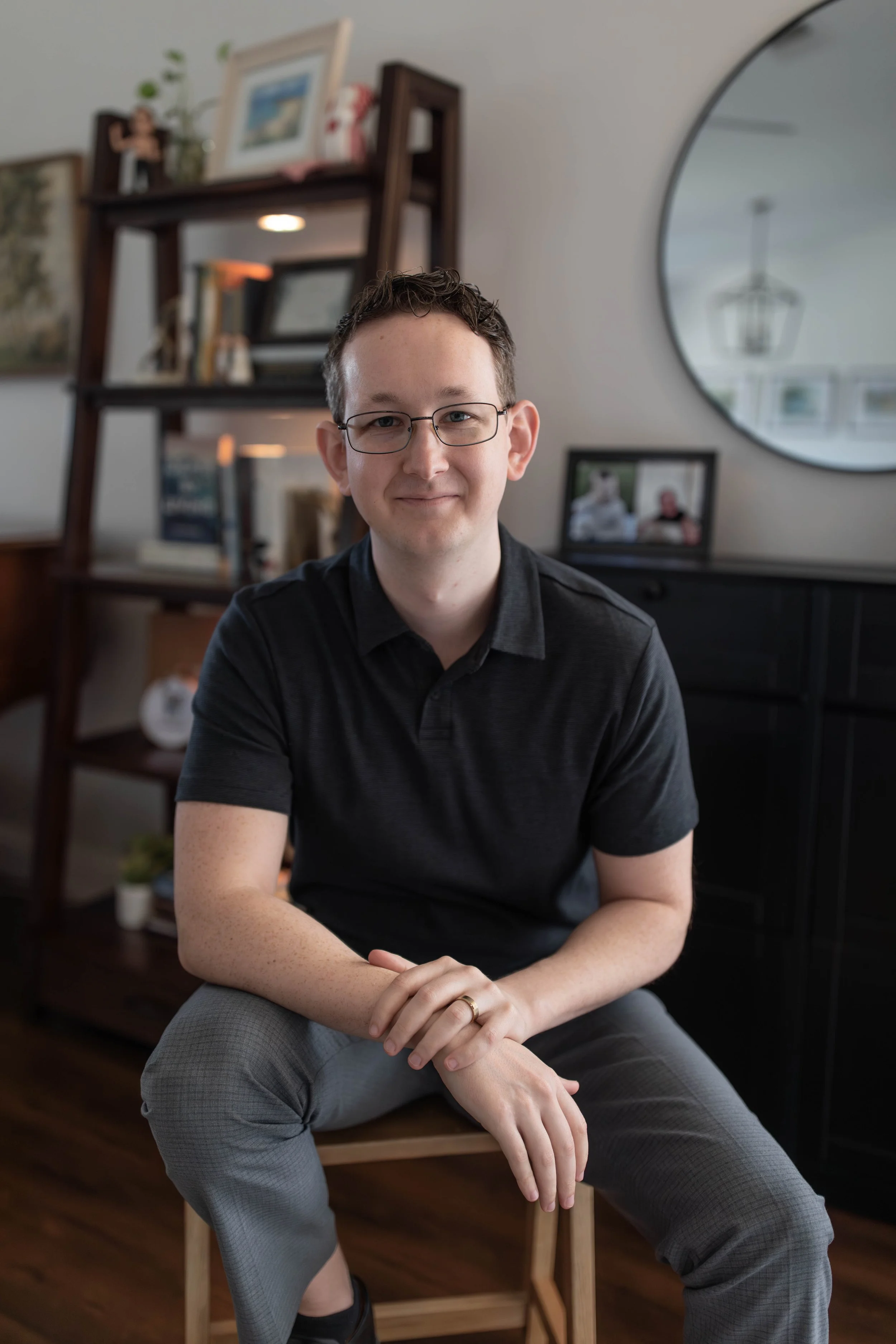 A young man with glasses, dressed in a black polo shirt and gray pants, sitting on a wooden chair in a living room. In the background, there is a dark wood bookshelf with framed photos and books, a round mirror on the wall, and a framed picture on a side table. He is smiling gently and looking at the camera.