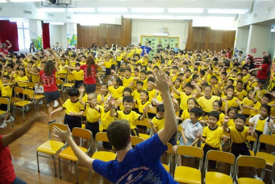 Large group of young children in yellow shirts participating in an activity in a school auditorium, with teachers guiding them.