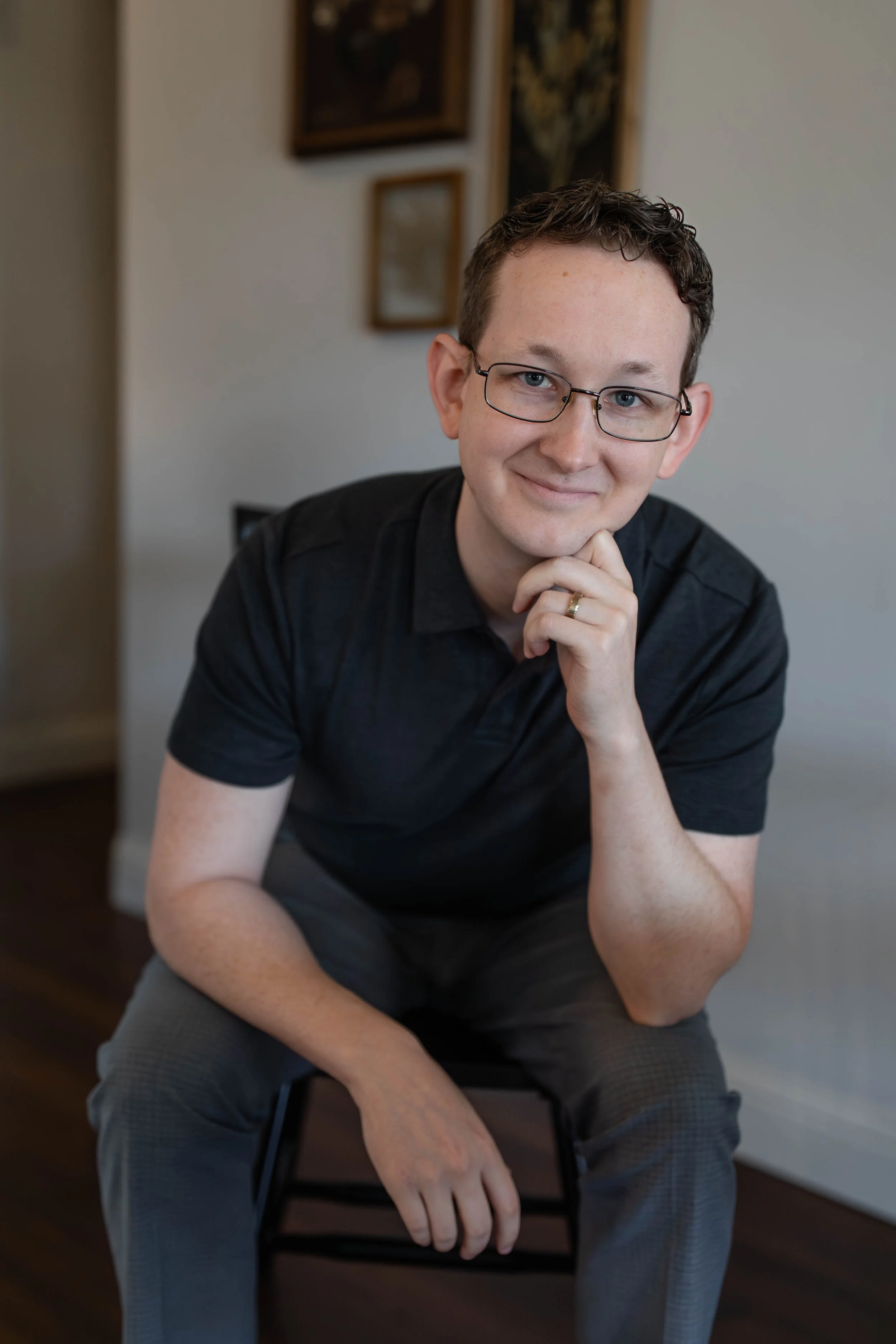 A young man with glasses and a wedding band sitting on a chair indoors, looking at the camera with a gentle smile, resting his chin on his hand.