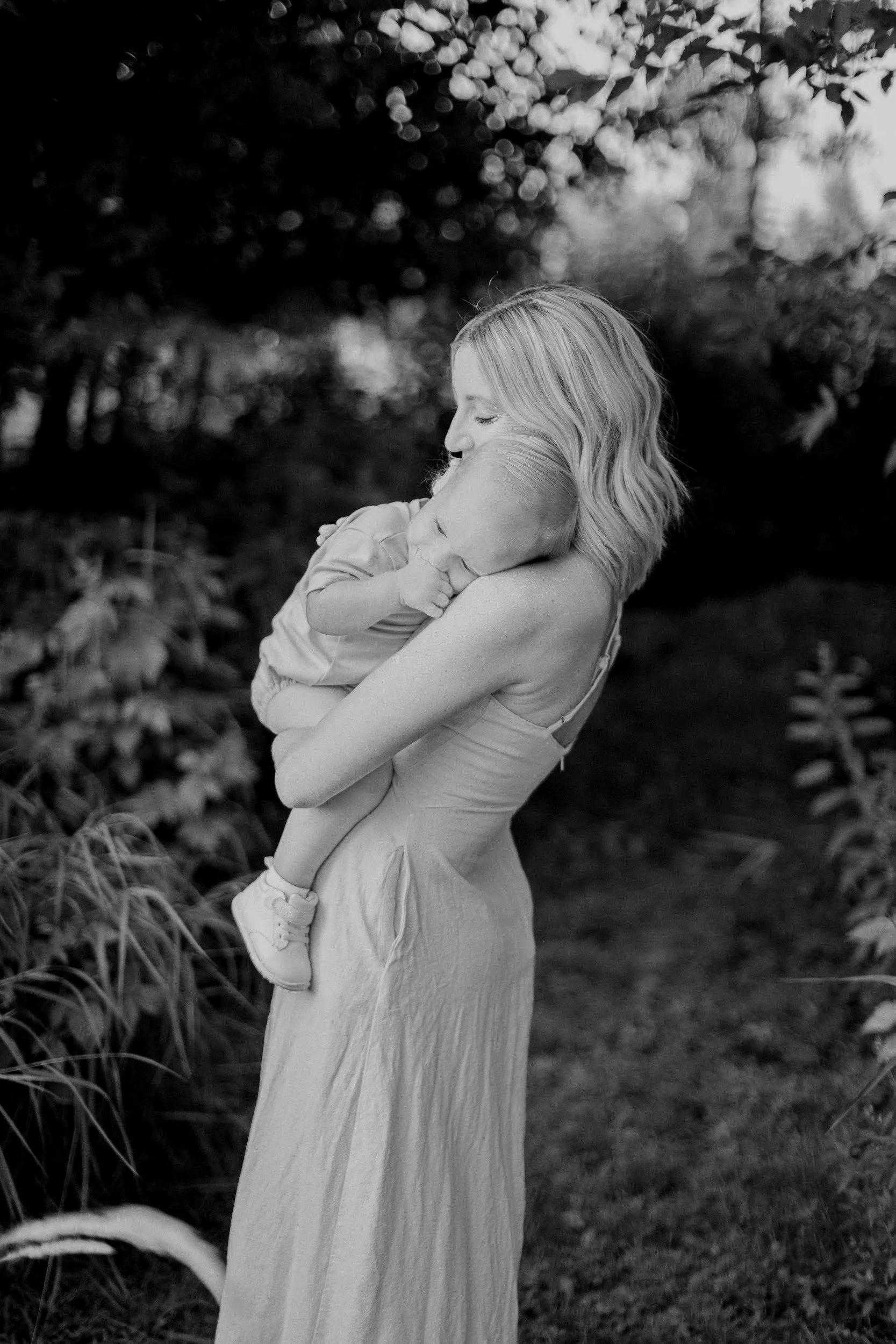 A woman with shoulder-length hair holds a sleeping young child in an outdoor setting with trees and foliage in the background, captured in black and white.