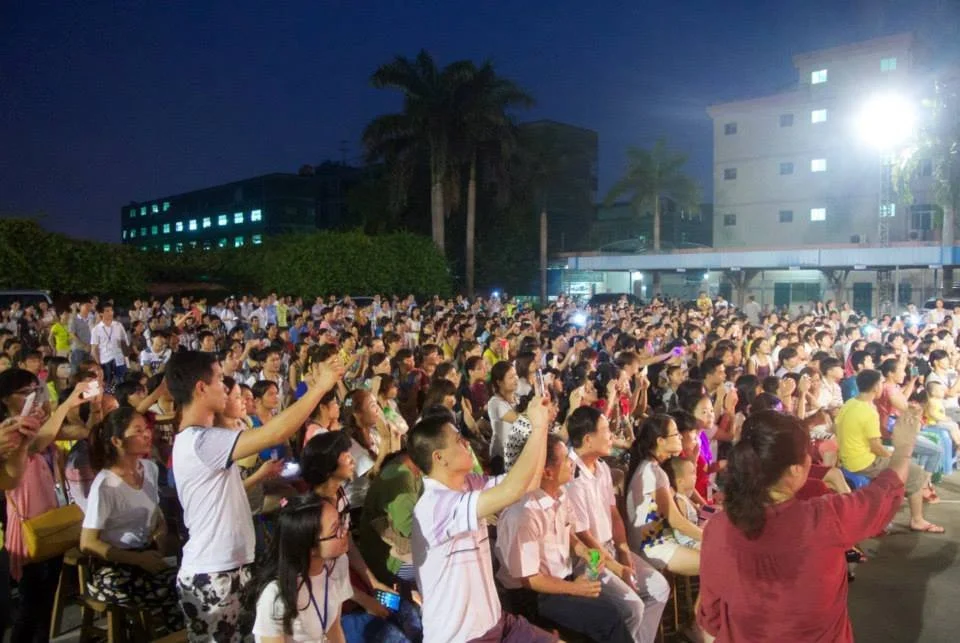 Large crowd of people seated outdoors at night, many holding up phones to record or take photos, with trees and buildings in the background.