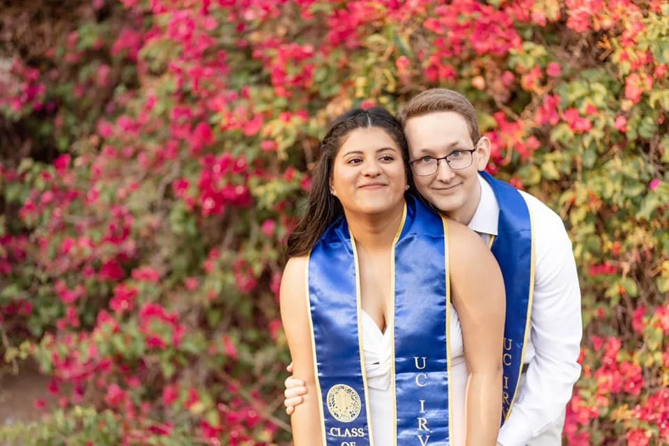 Two graduates, a woman and a man, in caps and gowns with blue stoles, standing outdoors in front of a bush with pink flowers, smiling and posing for a photo.