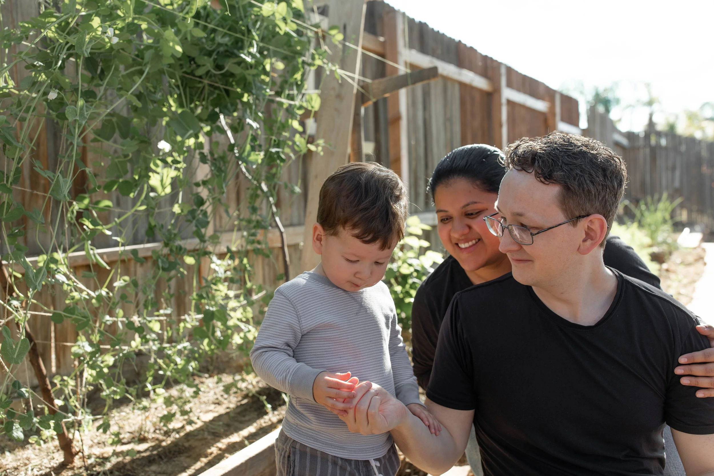 A family of three, including a young boy with brown hair, a woman, and a man with glasses, smiling outside in a garden with plants and a wooden fence.