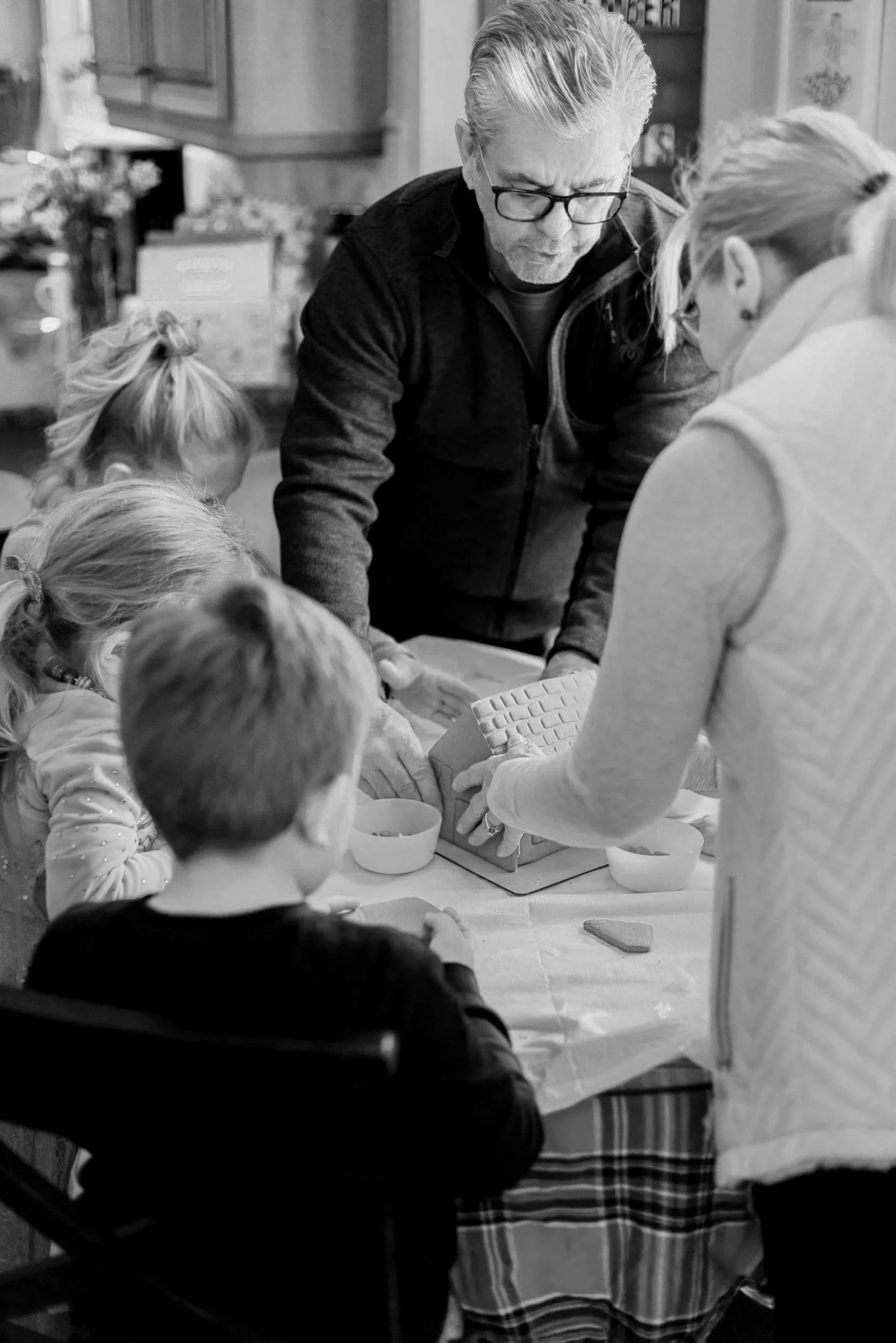 A man with glasses and a dark jacket is helping children decorate cookies at a table, with a woman assisting. The scene takes place in a cozy kitchen setting.