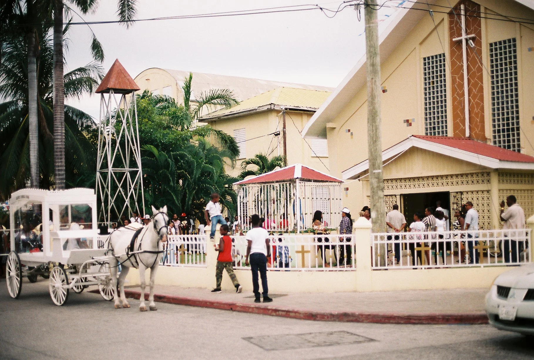 People gathering outside a church with a white fence, a horse-drawn carriage, and a small cage, with palm trees and buildings in the background.