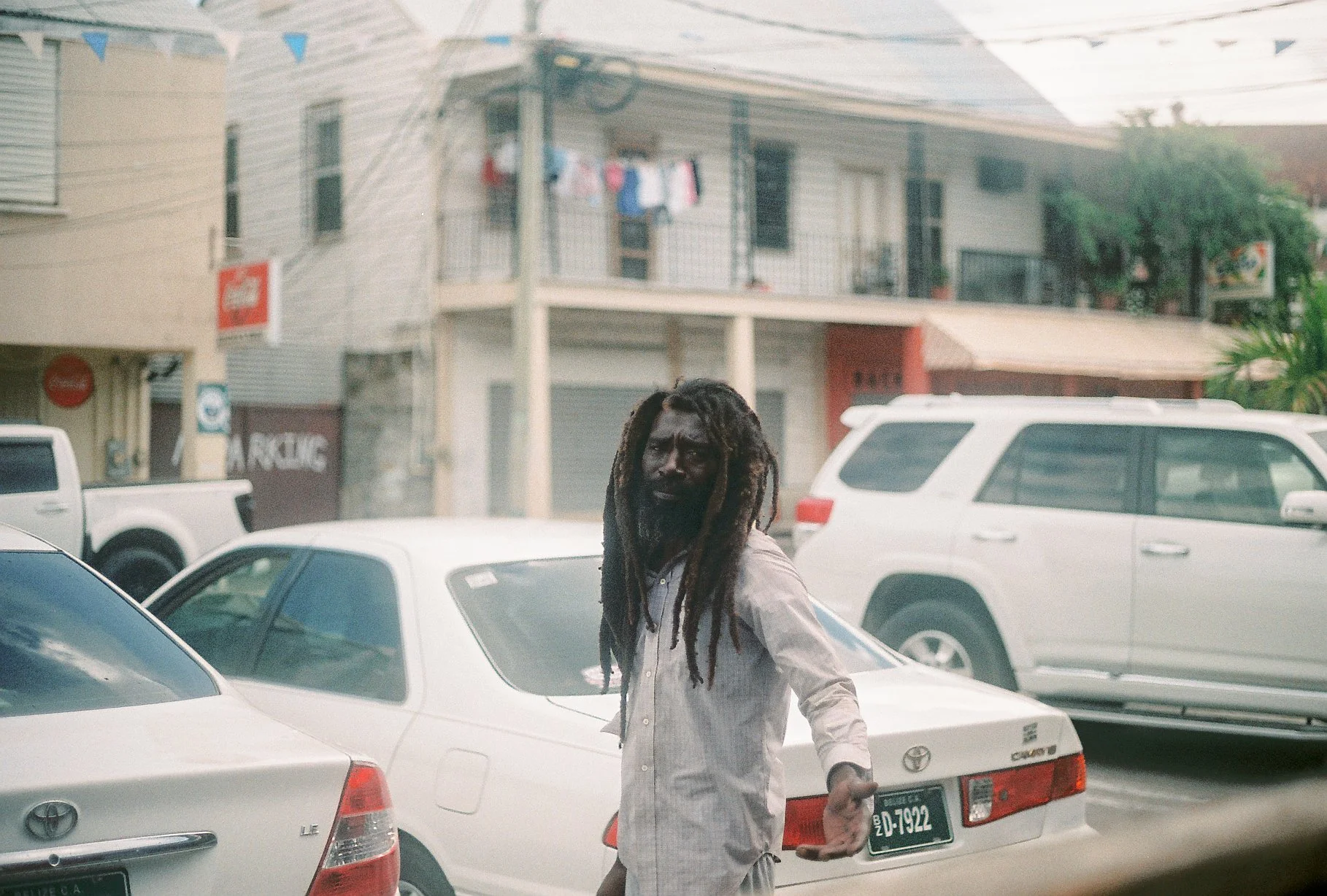 A man with dreadlocks and a beard wearing a light-colored shirt, standing in front of a white Toyota car in a parking lot with other vehicles, with apartment buildings in the background.