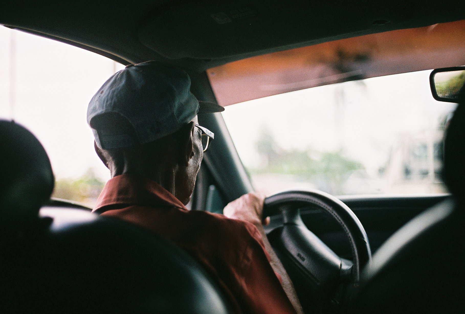 An elderly man wearing a cap and glasses driving a vehicle, seen from the backseat, with an out-of-focus landscape through the windshield.