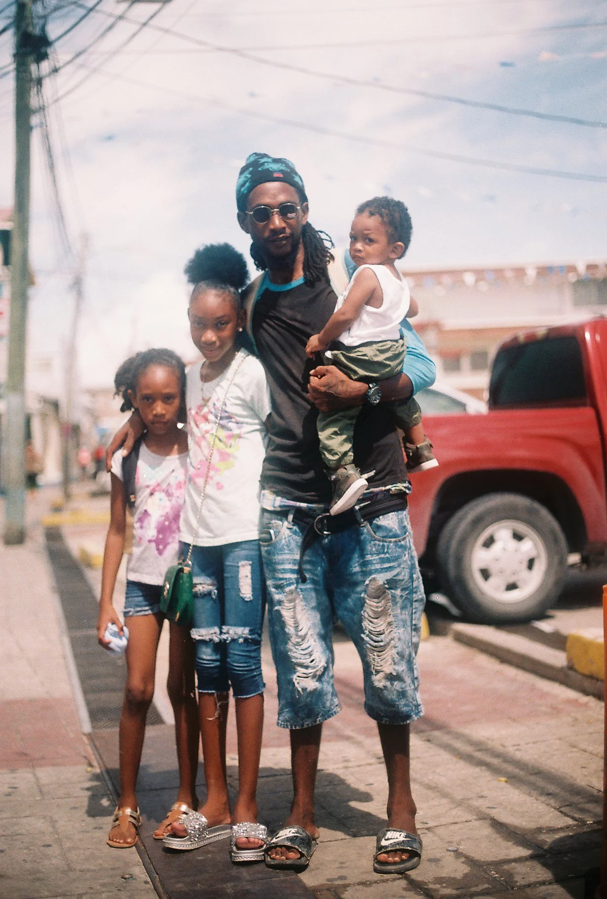 A man holding a young boy in his arms, standing on a sidewalk with three young girls. The man is wearing sunglasses, a hat, and ripped jeans. The background shows a street scene with a red truck and power lines.
