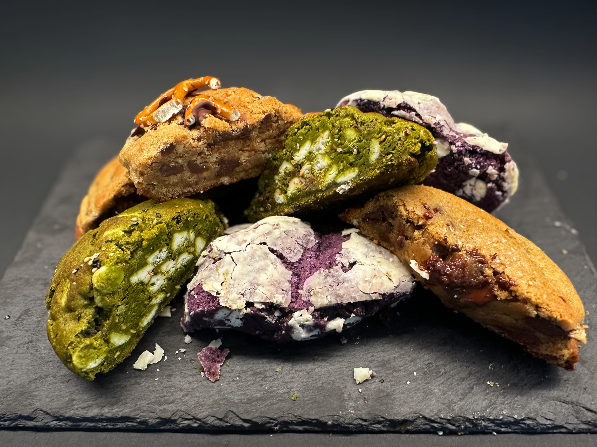 Blunderbuss Baking Co. small batch cookies — matcha white chocolate, ube, bourbon espresso, and miso pretzel, cut open on a black slate board.