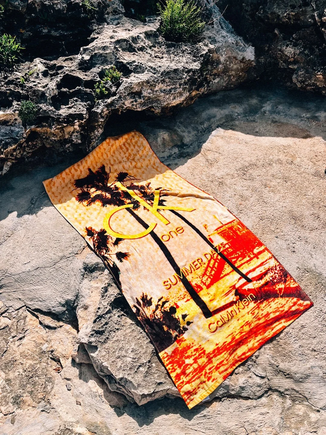 Calvin Klein towel with sunset and palm tree design lying on rocks. Designed by British Artist, Harry Cartwright