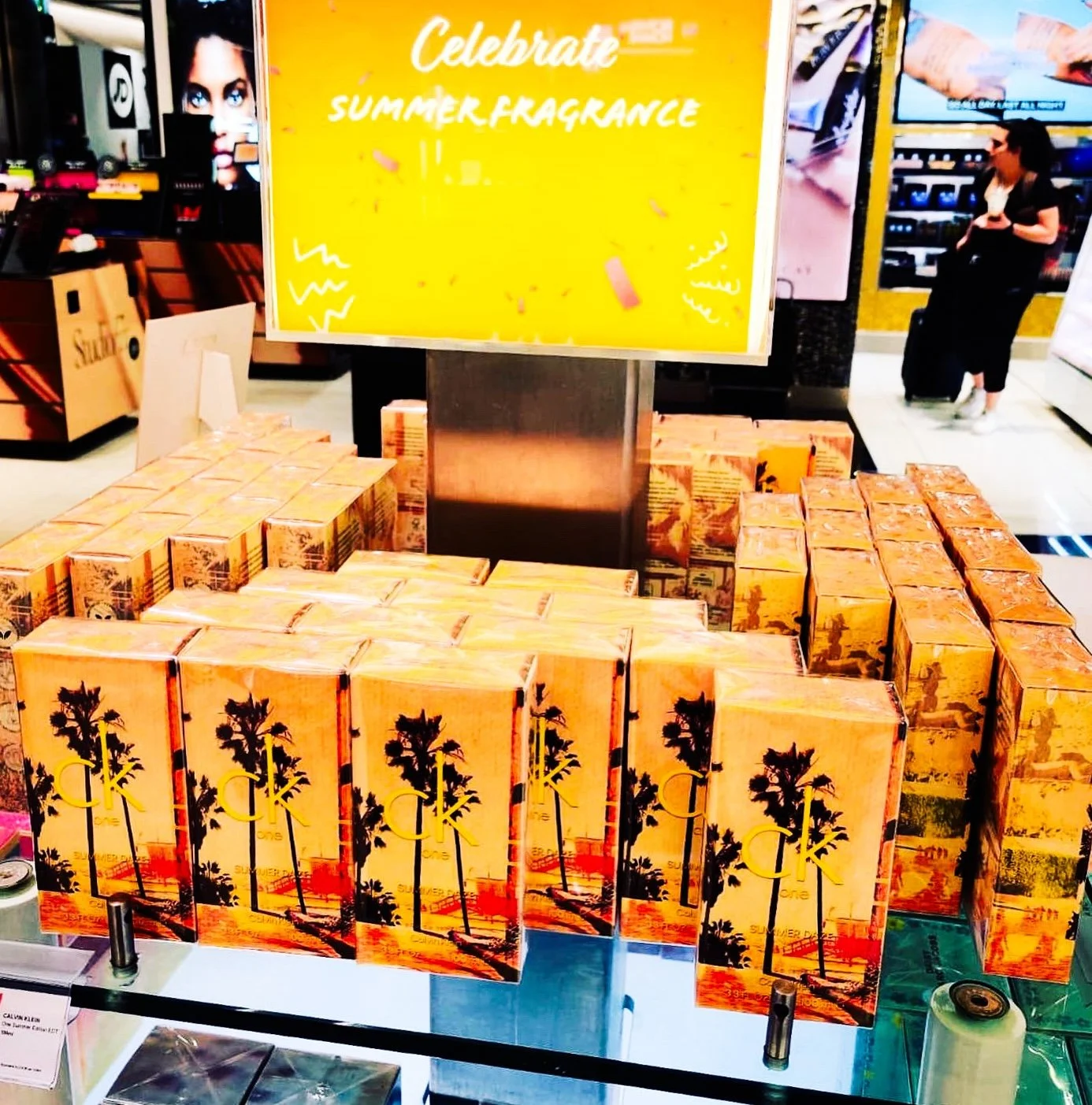 Display of CK One Summer fragrance boxes at a retail store with a yellow sign reading "Celebrate Summer Fragrance".