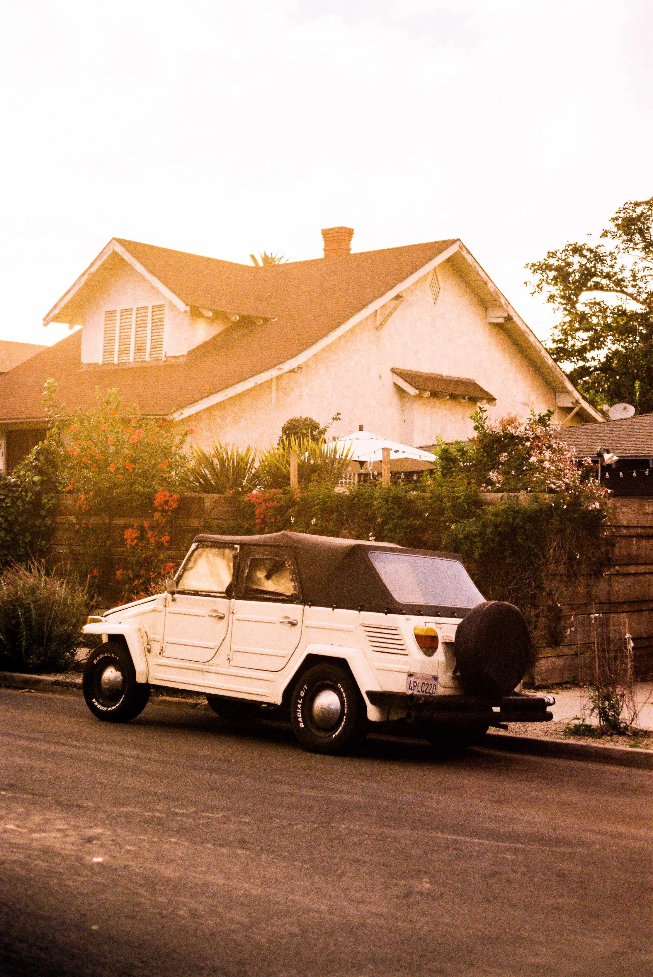 A white vintage car with a black soft top parked on a residential street during sunset, with a house, garden, and trees in the background.