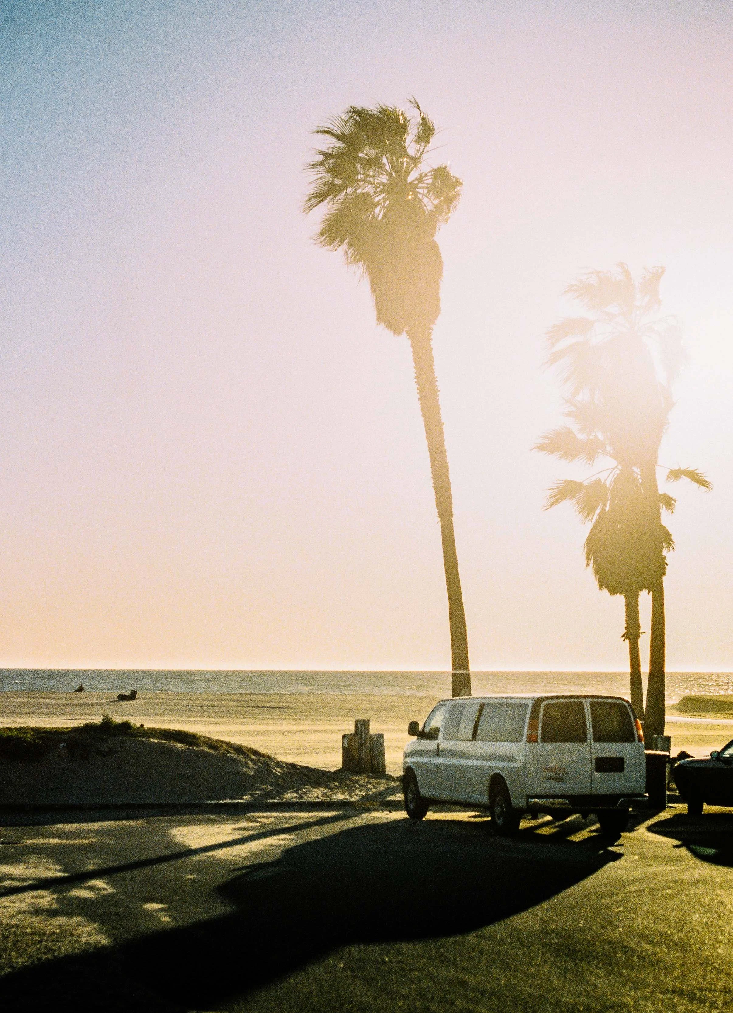 Sunset at the beach with two palm trees, a white van, and shadows on the ground.