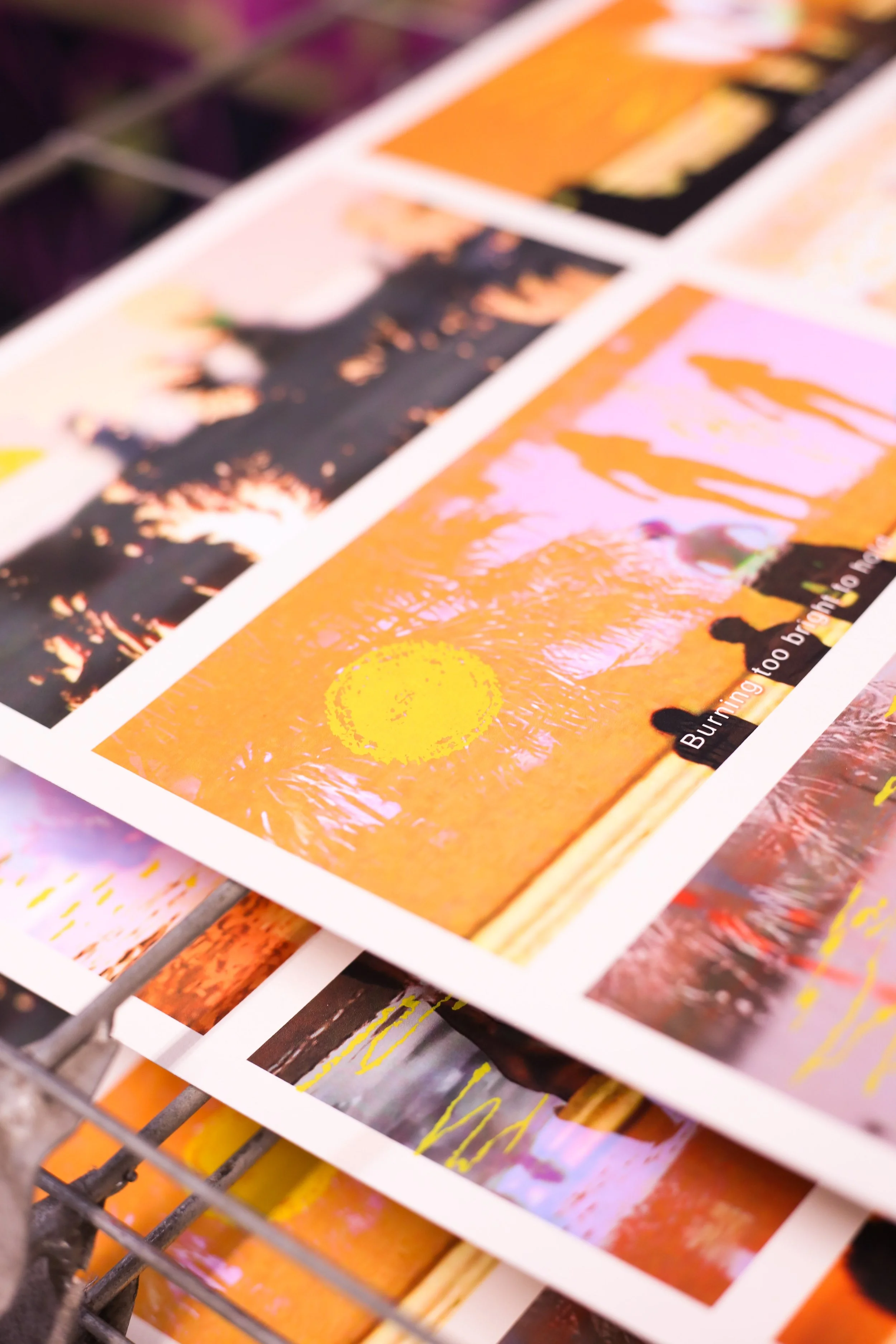 Photograph of printed photographs with vibrant sunset and landscape scenes, some silhouettes of people, and an orange and pink color palette, stacked on a metal rack.