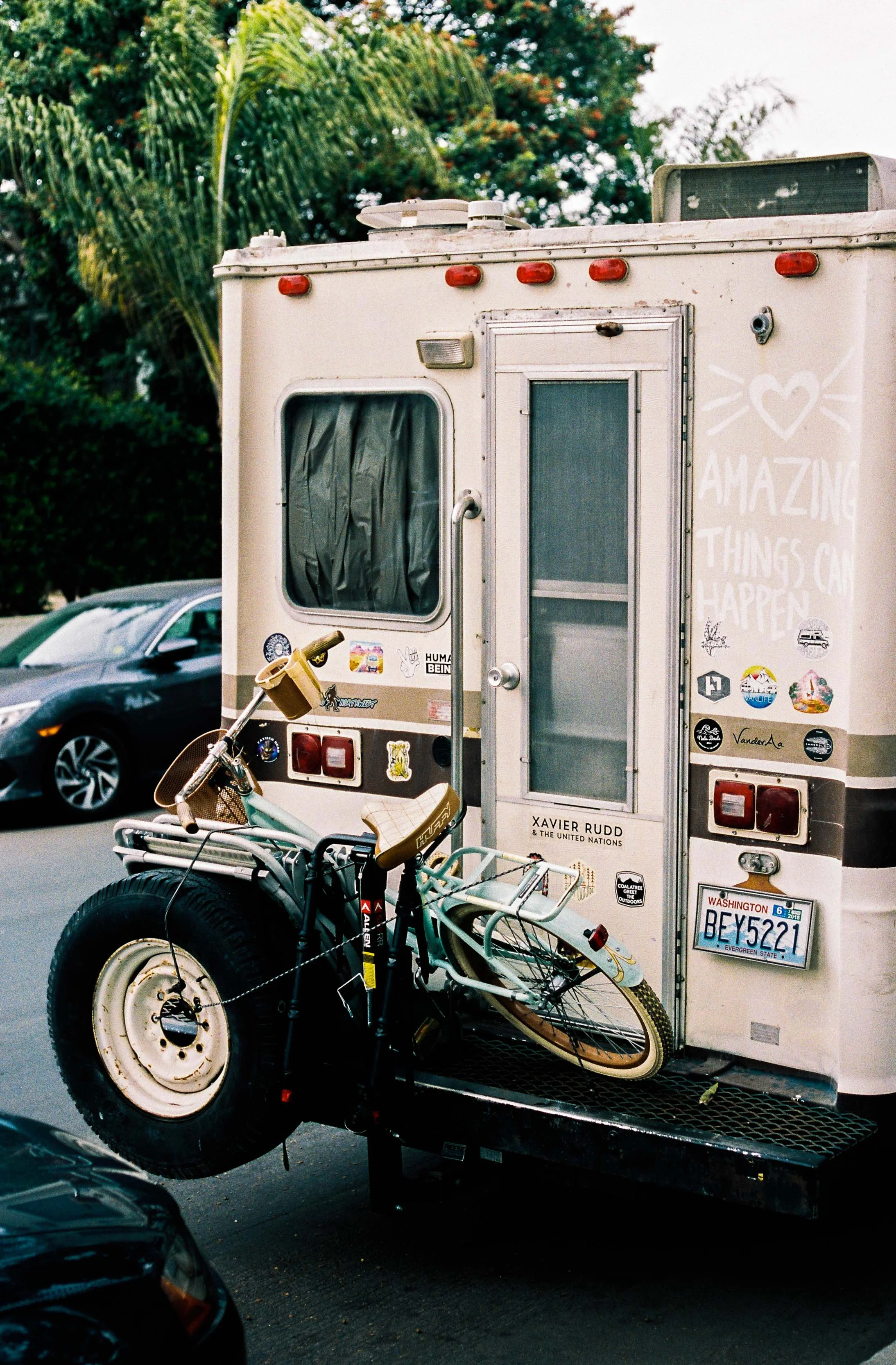 A vintage camper trailer with stickers, a bicycle mounted at the back, and a message that says "Amazing Things Can Happen" on the side, parked on a street with trees in the background.