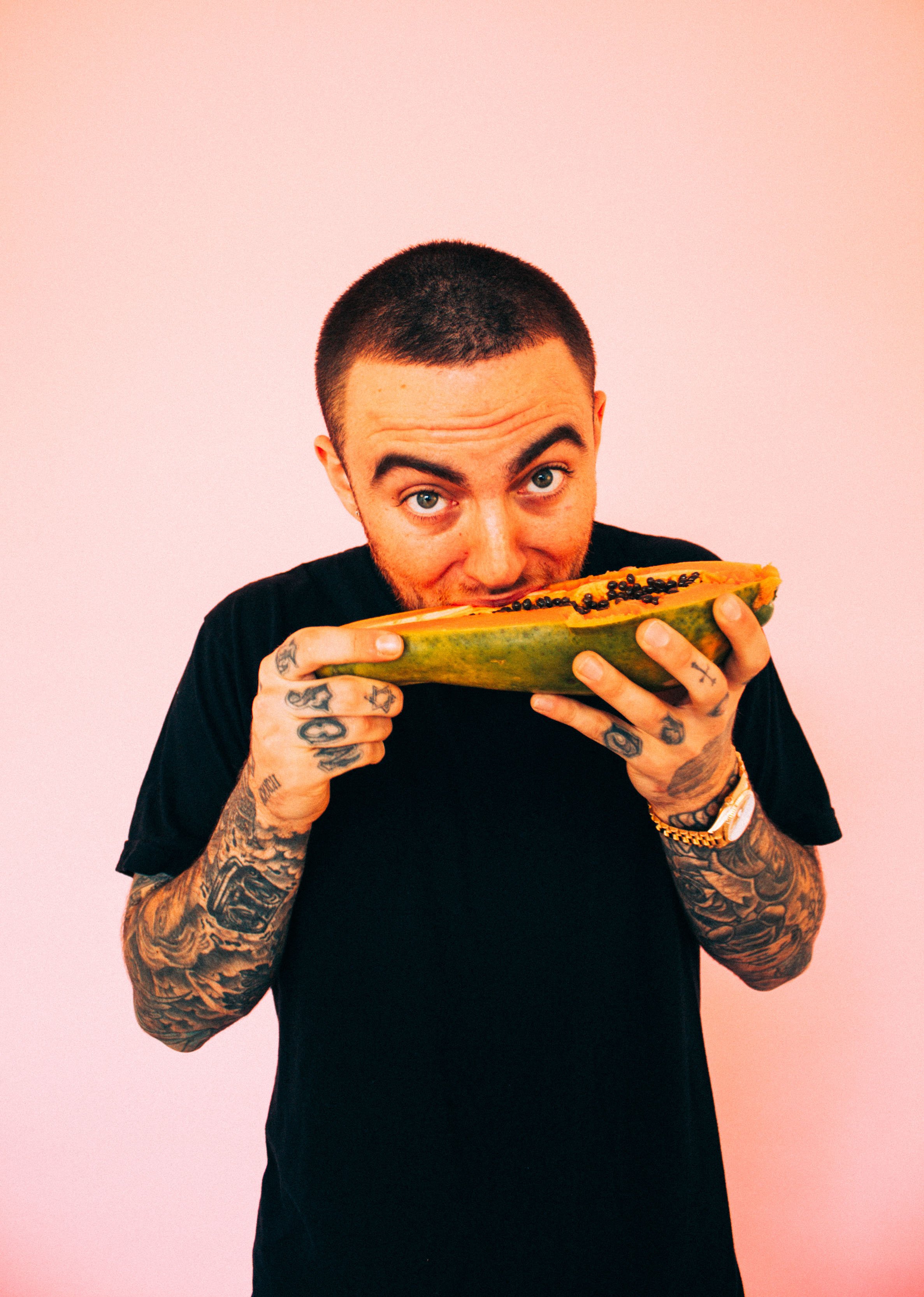 Mac Miller holding a halved papaya with black seeds and taking a bite from it against a plain light pink background. Photograph by British artist, Harry Cartwright.