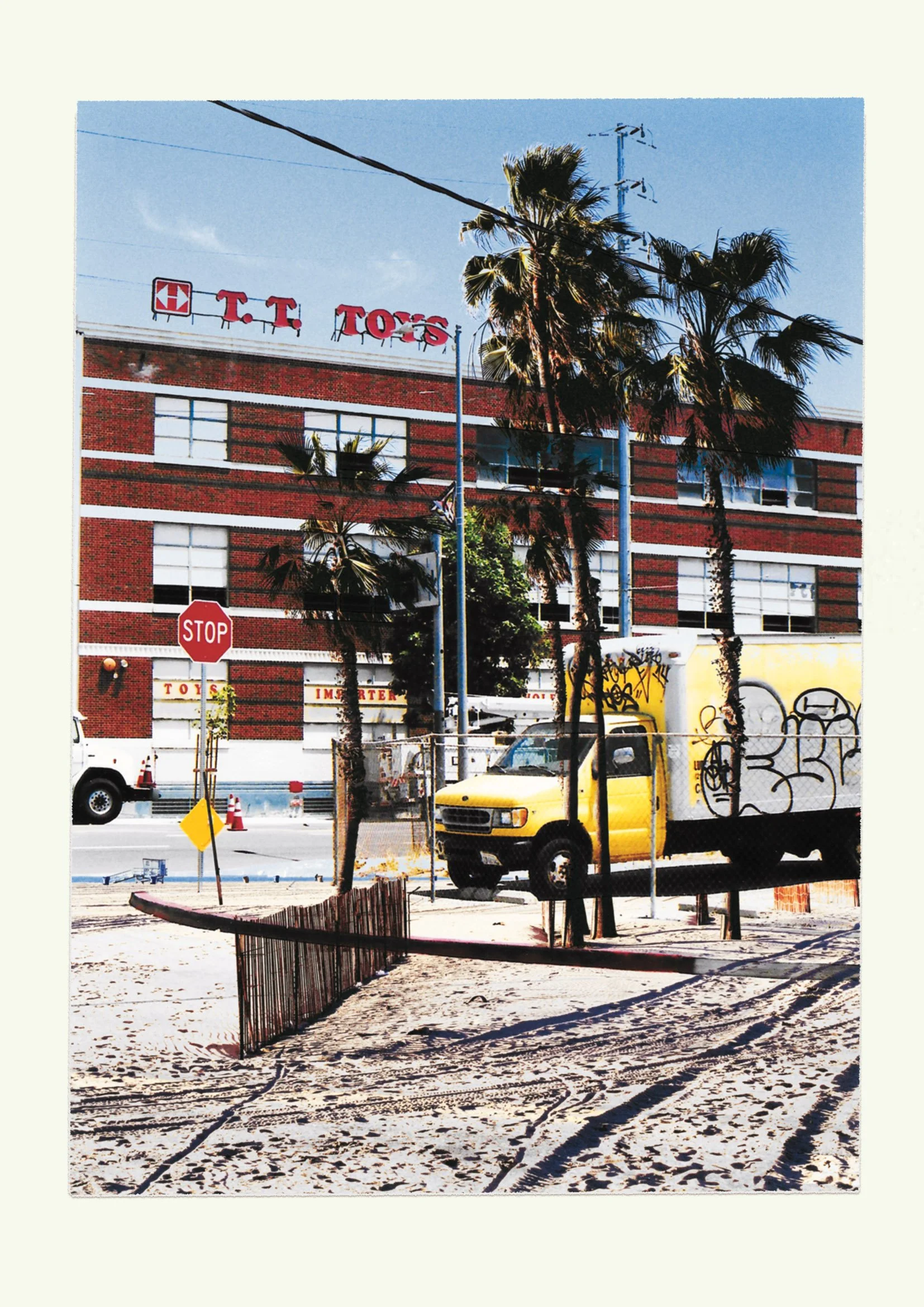 Street scene with palm trees, a yellow delivery truck with graffiti, a stop sign, and a large brick building with a T.T. TOYS sign on the roof.