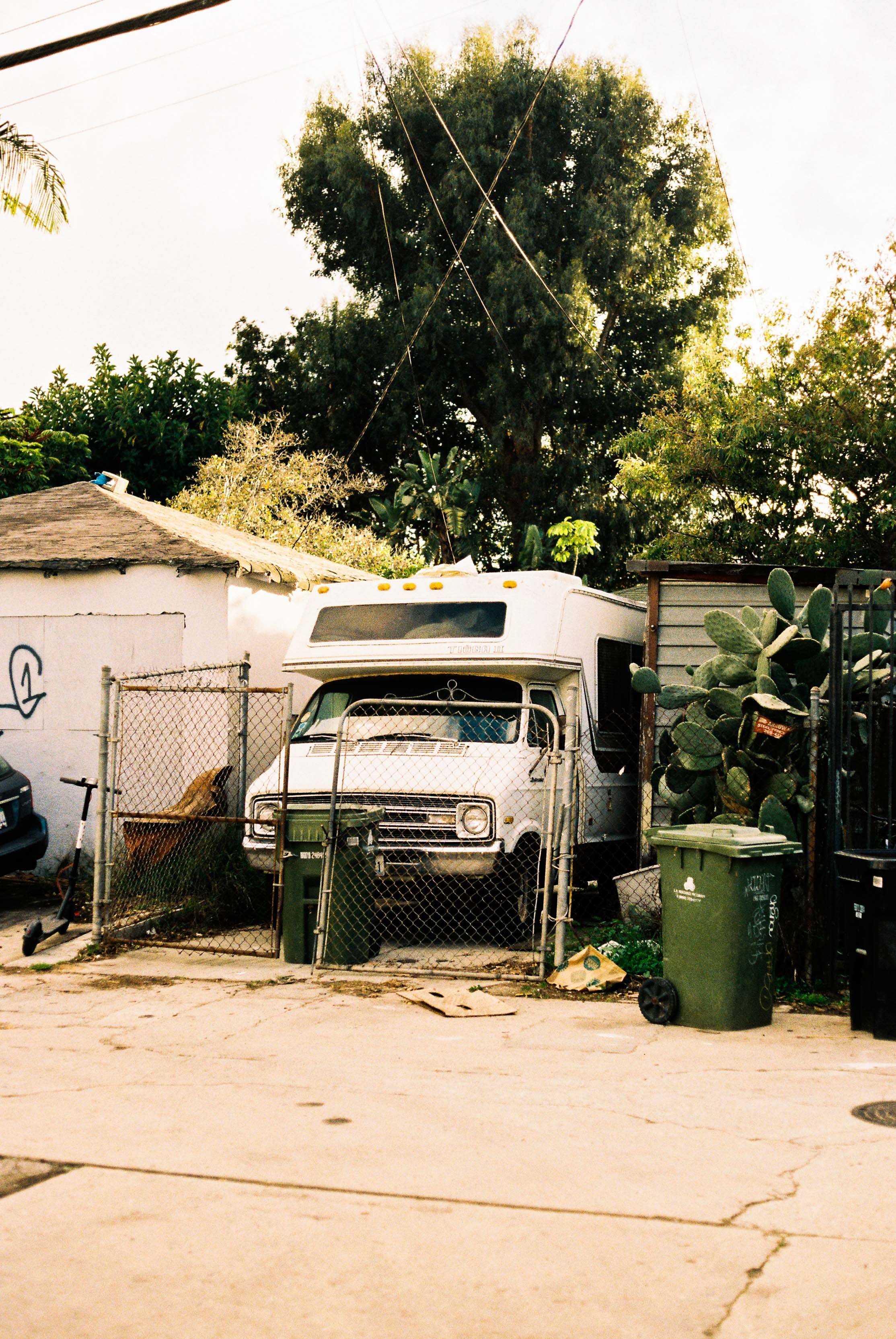 An old white camper van parked inside a fenced yard with surrounding green plants and trees. There are trash bins near the gate and a cactus plant on the right.