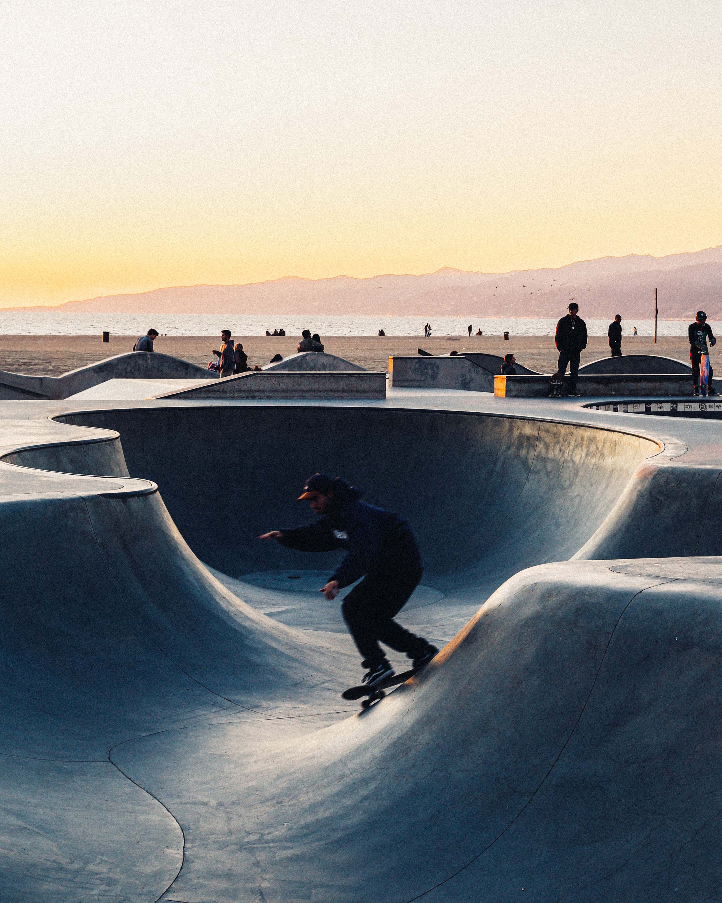 Skateboarder riding in a concrete skatepark near the beach during sunset with other people in the background.