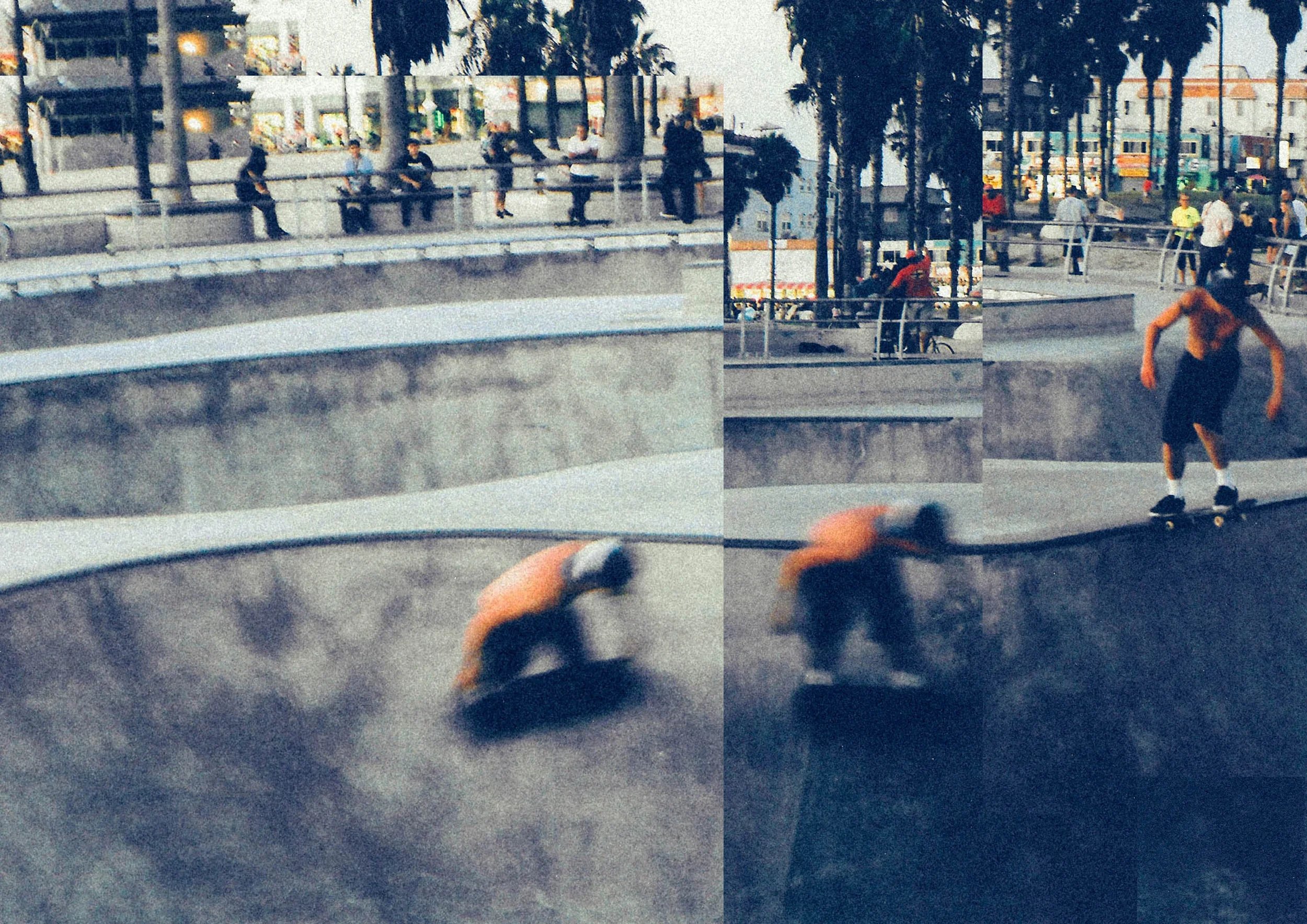 A skateboarder wearing an orange shirt and black shorts performing a trick on a skateboard at a skate park, with people sitting and walking in the background and trees lining the area.