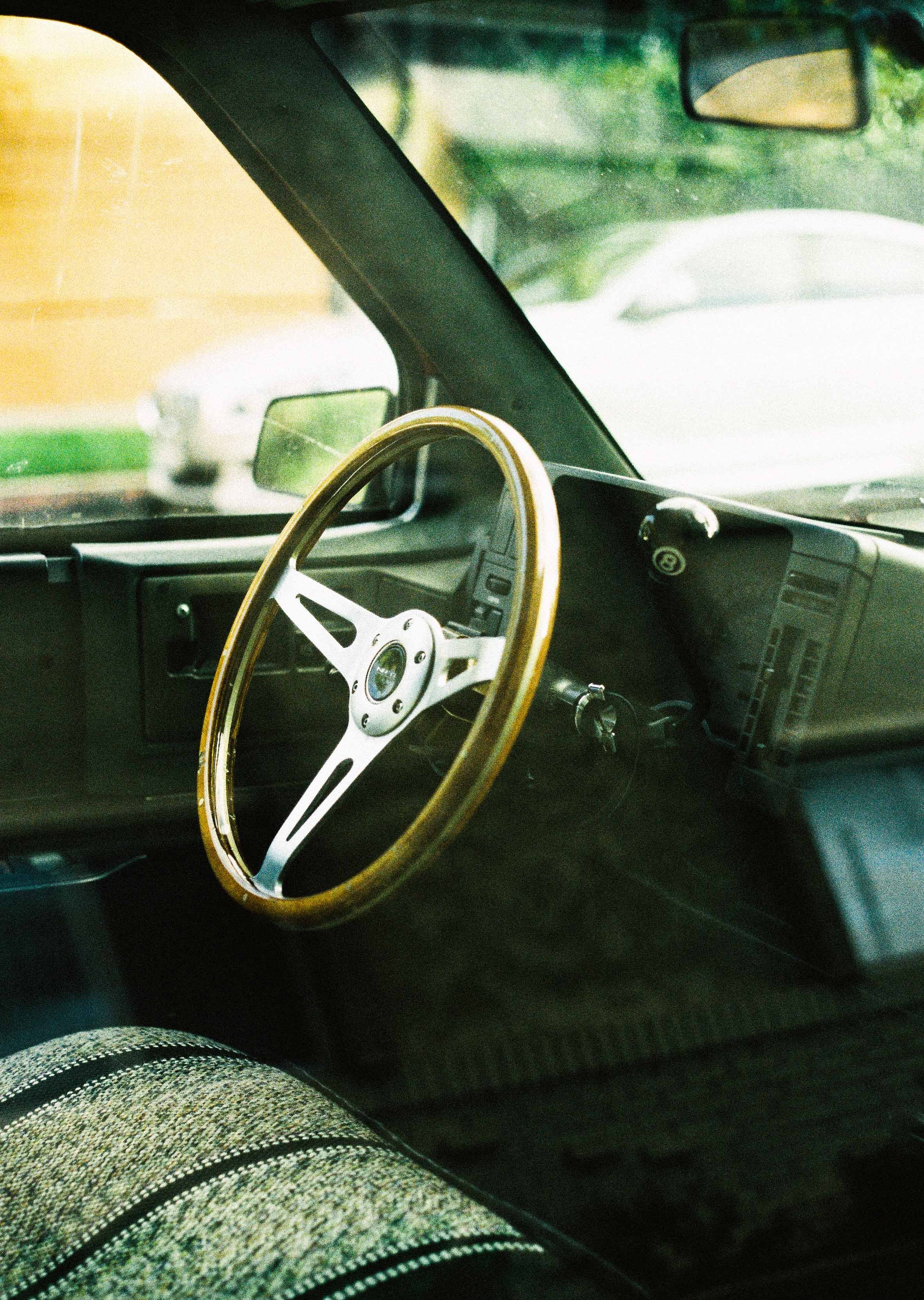 View of a vintage car interior, showcasing a wooden steering wheel, dashboard, and rearview mirror, with cars visible through the windshield.