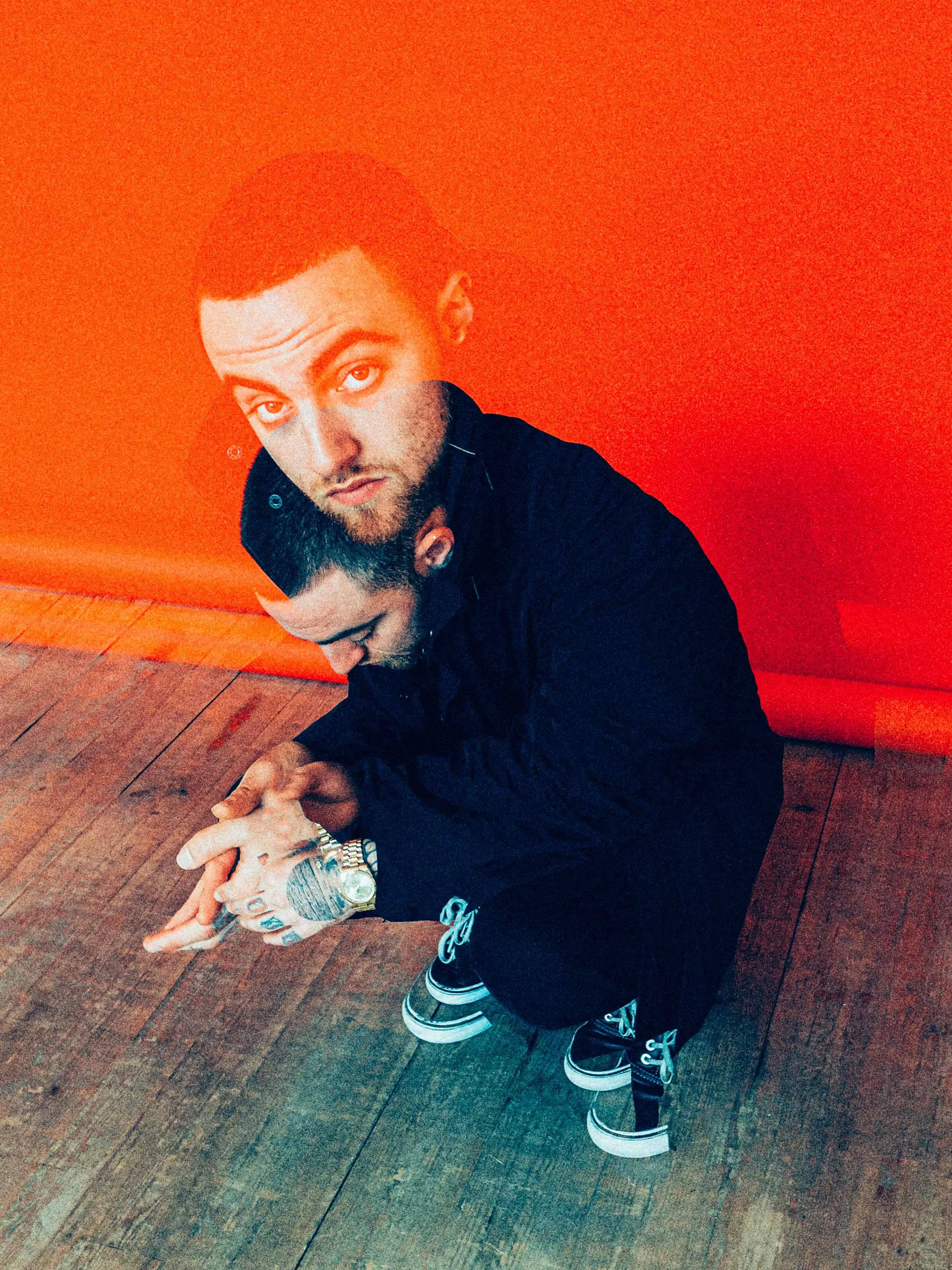 A young man is crouching on a wooden floor against a vibrant red wall, looking up at the camera with an intense expression. He is wearing a black jacket, sneakers, and has tattoos visible on his hands. The photo has a double exposure effect, merging the man's image with a semi-transparent overlay.
